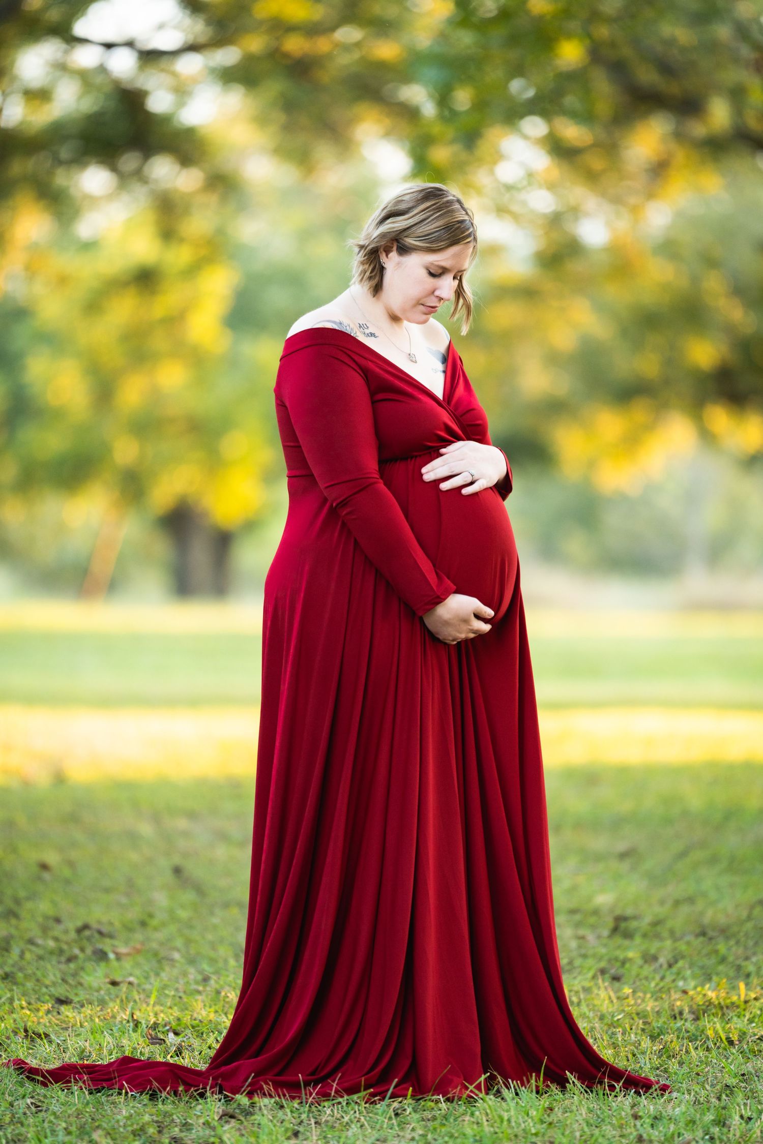 Expectant mother in a flowing red dress poses in a sunlit garden setting.