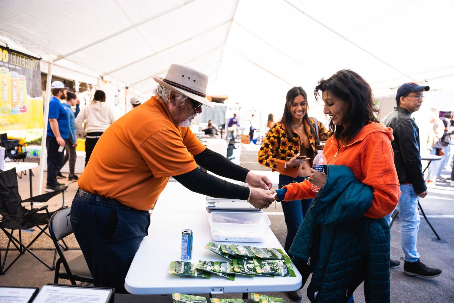 People interacting at a festival merchandise booth under white tent canopy.