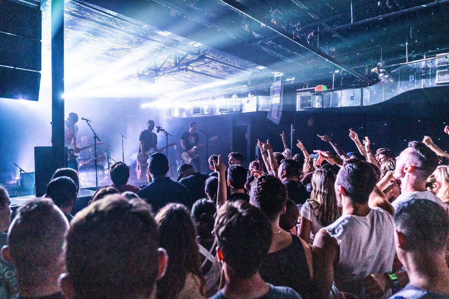 Enthusiastic crowd with raised hands enjoying a live concert performance in a dimly lit venue.
