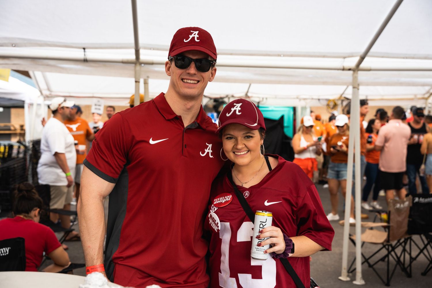 Two fans in crimson athletic gear pose together under a white tent at a sports event.