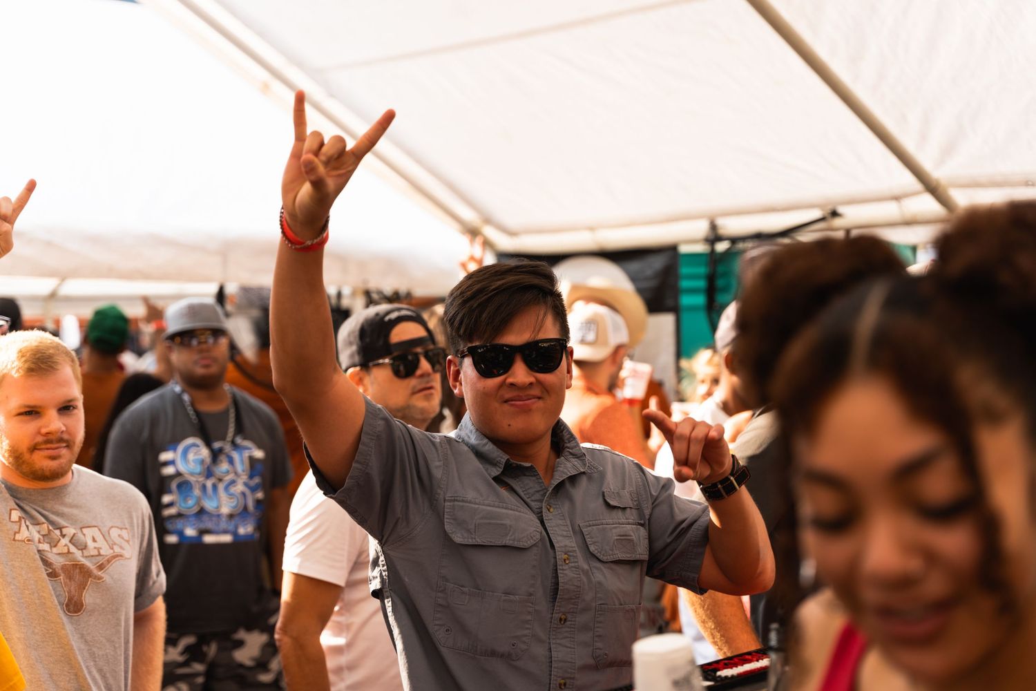 Enthusiastic crowd makes rock hand gestures under a white tent at an outdoor event.