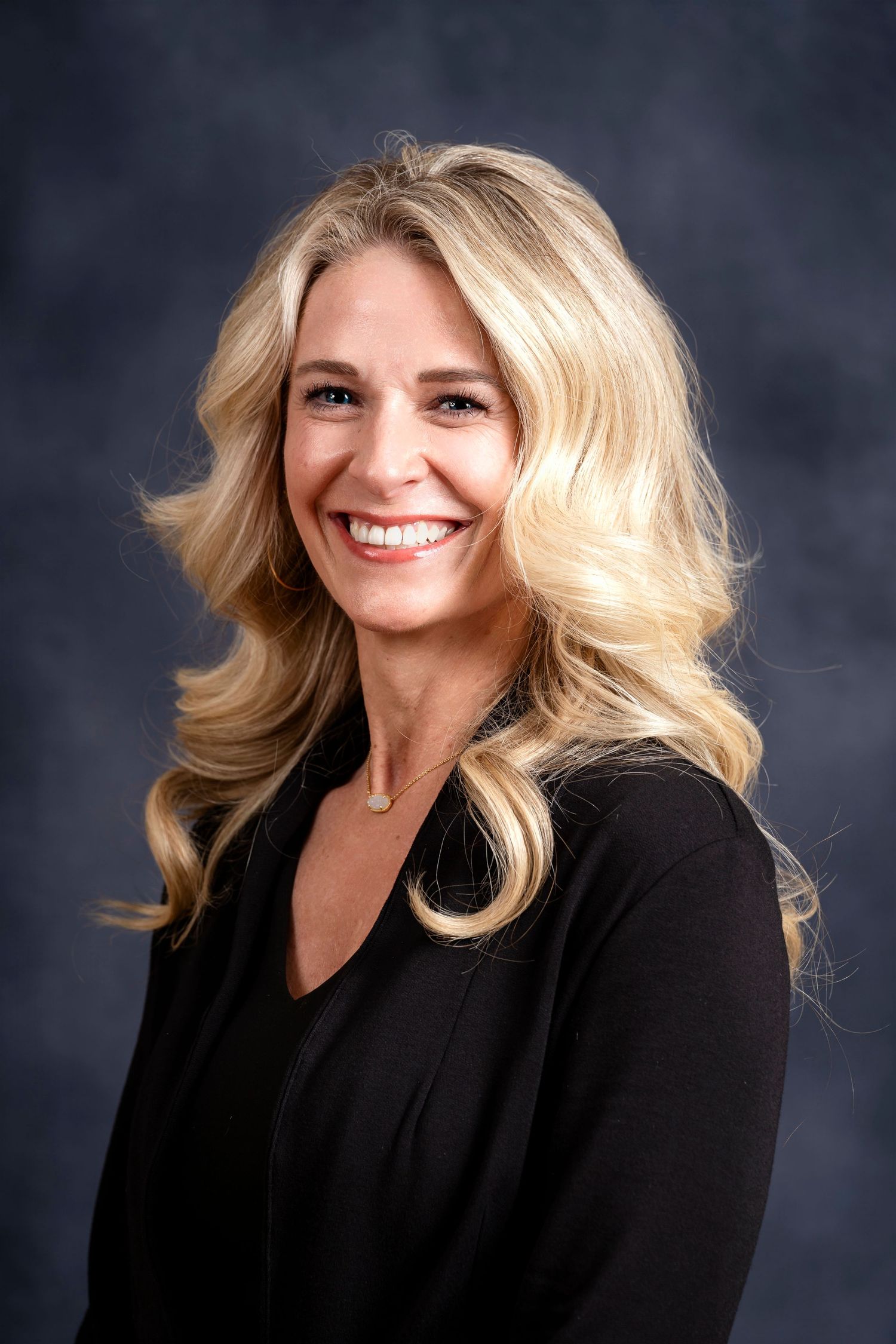 Side-angled professional portrait of a person with blonde hair wearing a black blazer against a dark backdrop.