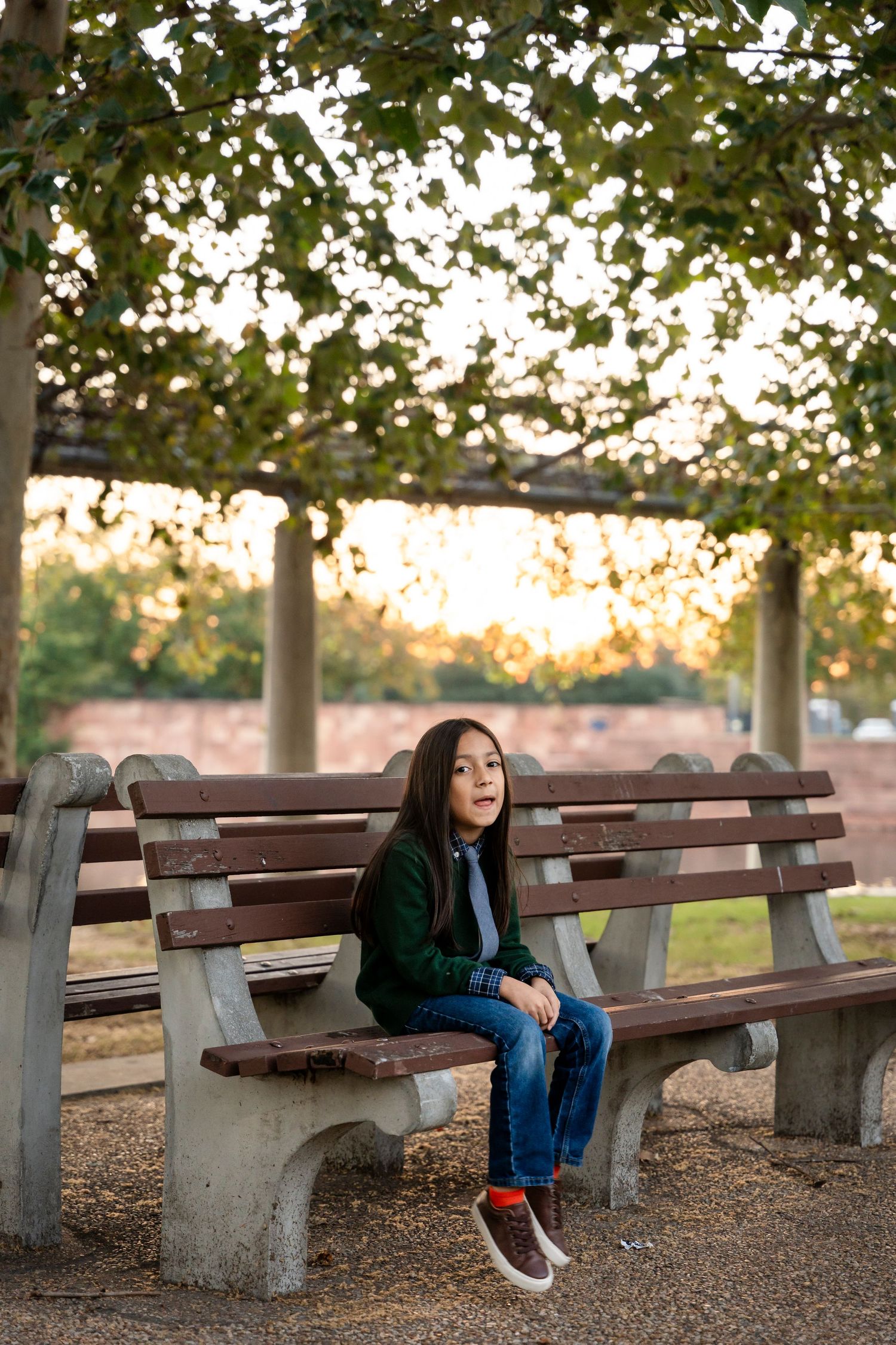 Young person in green sweater sits alone on park bench at sunset.
