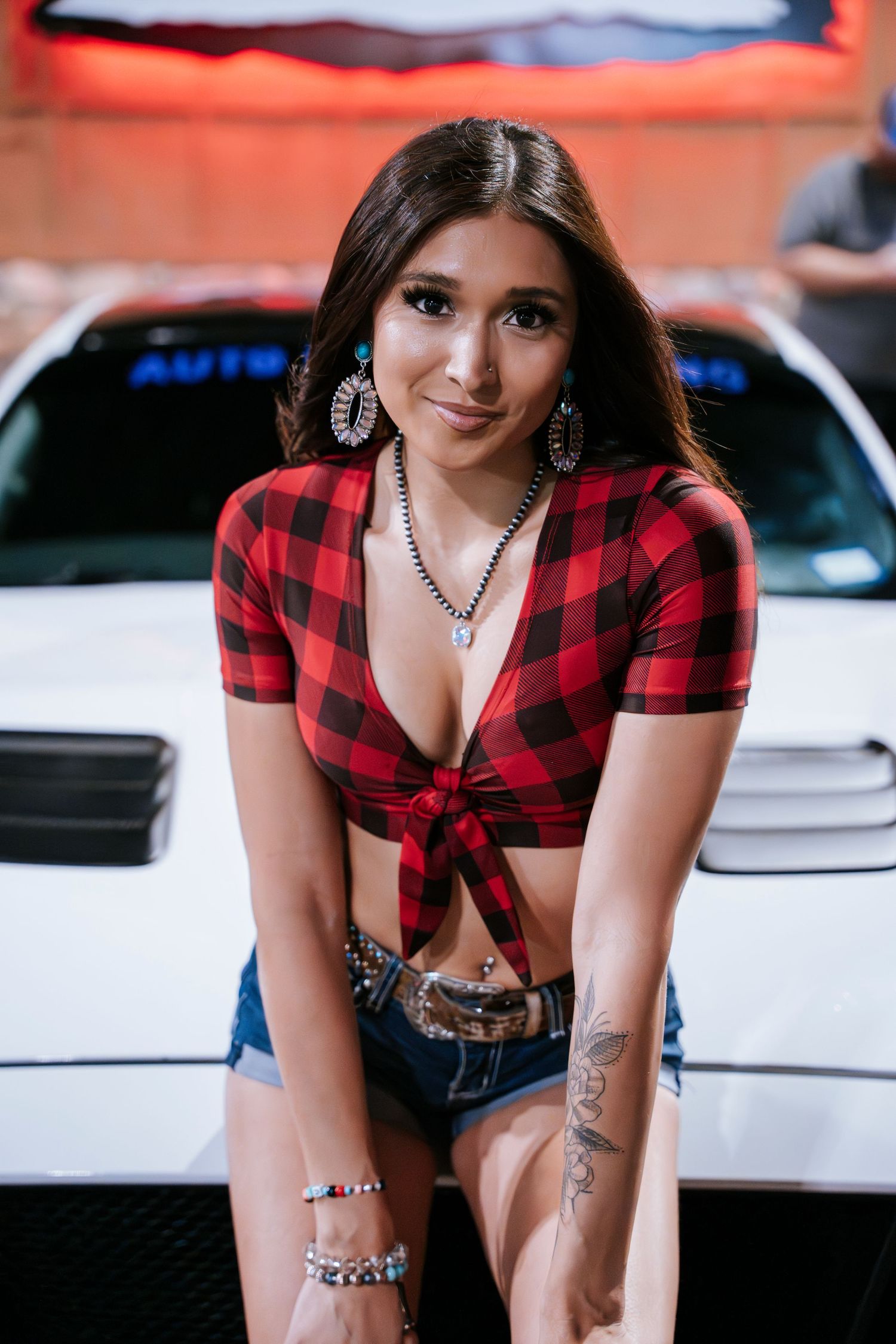 Woman in buffalo plaid top and jewelry poses by white car at night event.