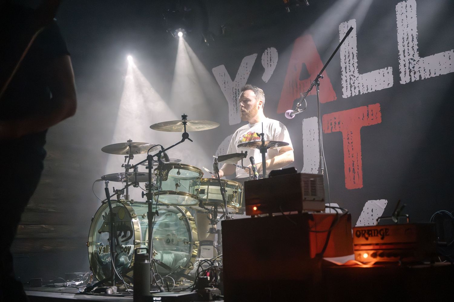 A drummer performs on stage with dramatic lighting and smoke effects at a concert venue.