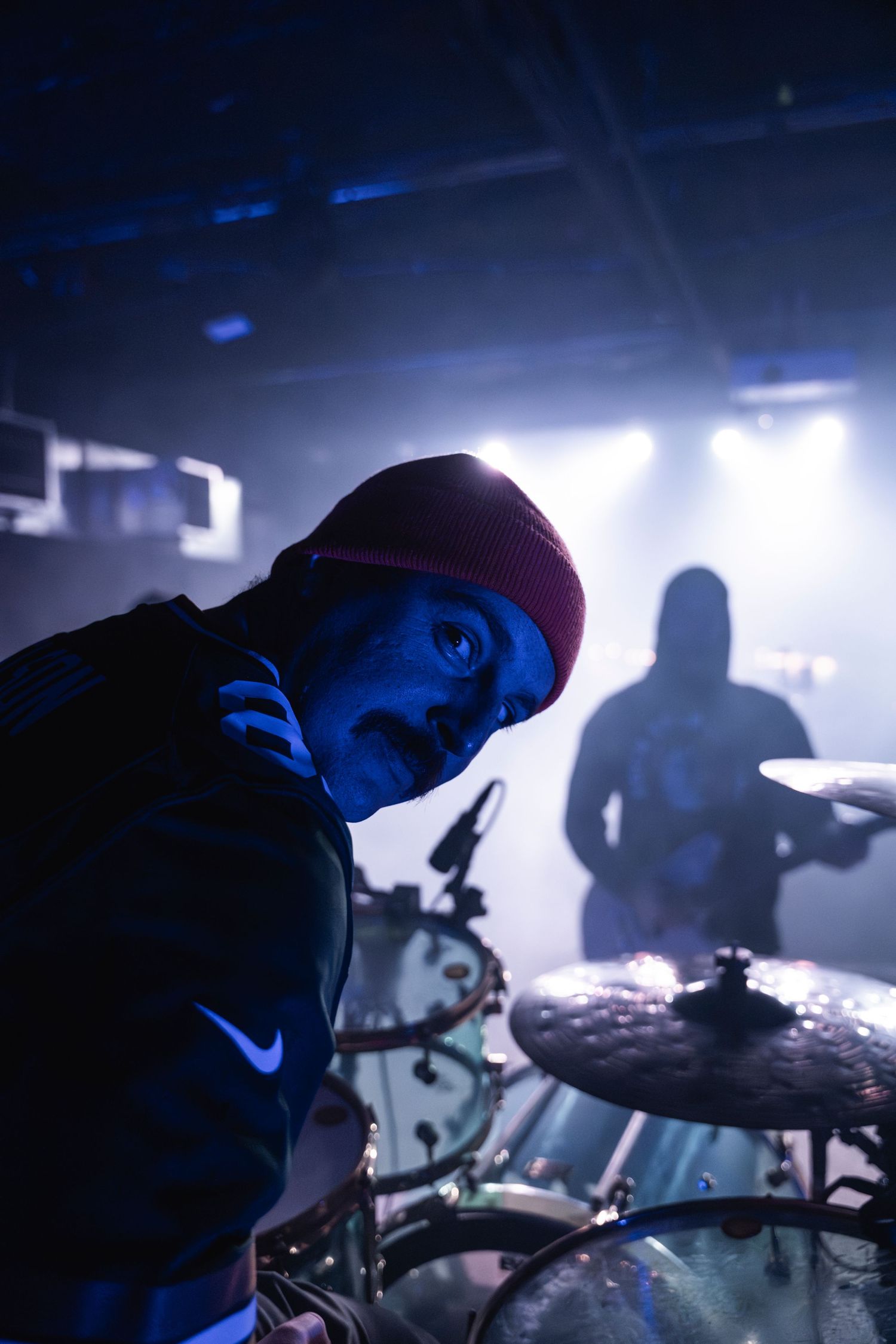 Drummer in beanie performing under blue stage lighting with dramatic shadows.