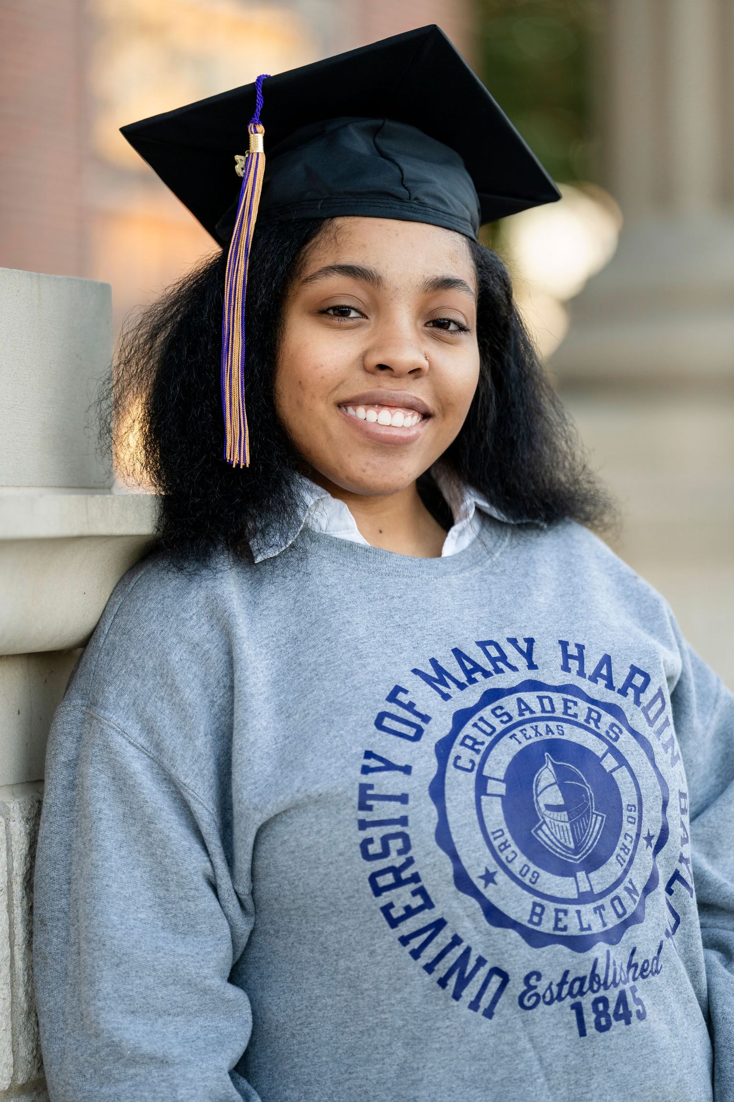 A graduate wearing a black mortarboard poses in a gray university sweatshirt against a columned background.