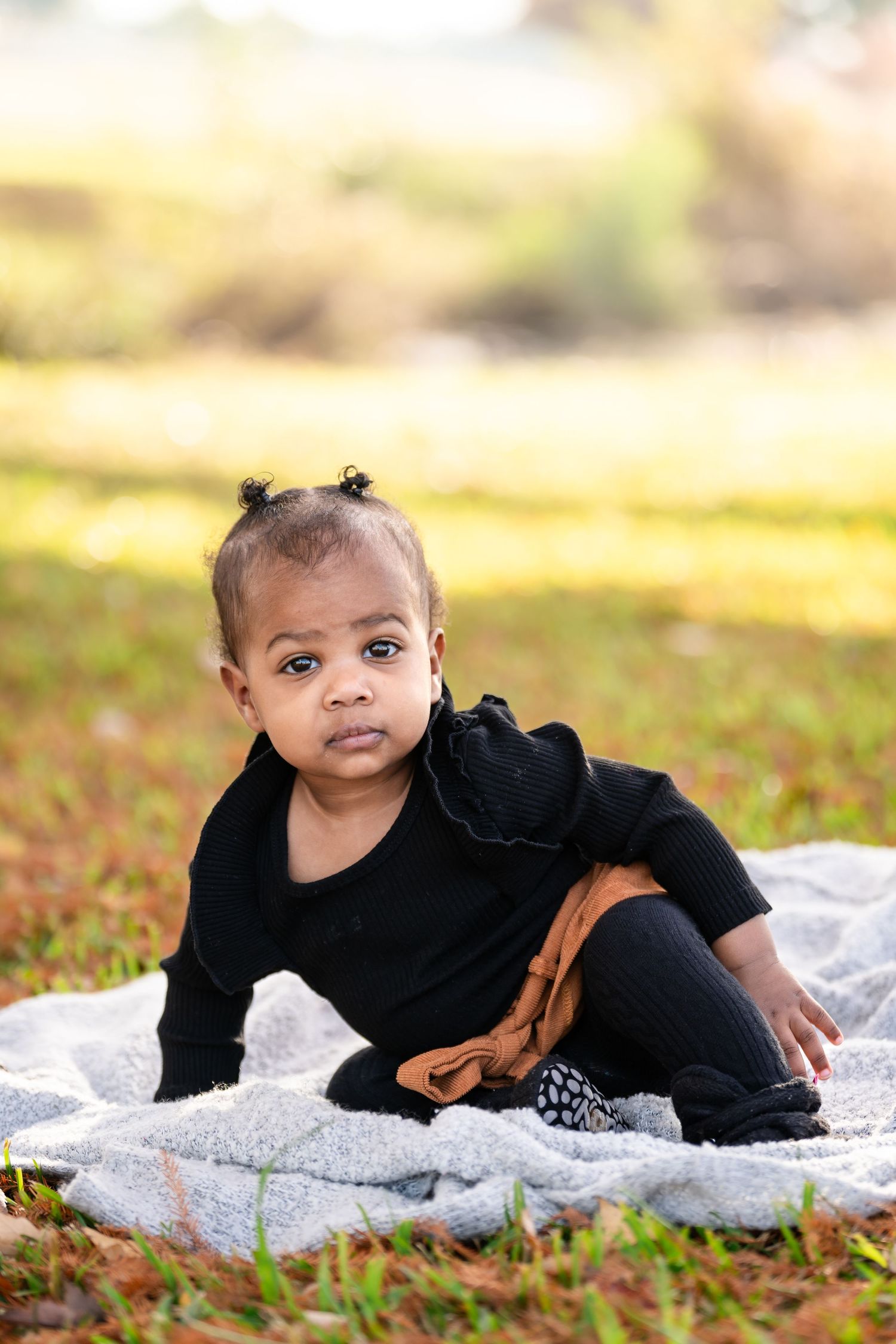 Baby in black outfit sits on blanket during outdoor autumn photo session.