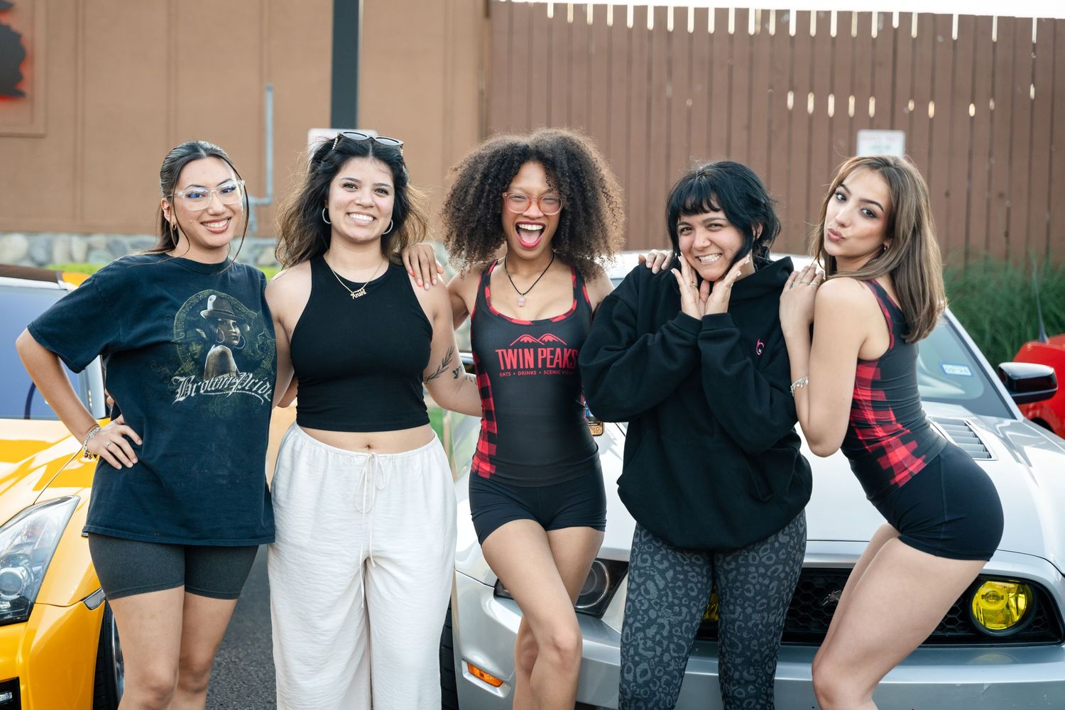 Group of friends posing together in casual outfits against a car in a parking lot.