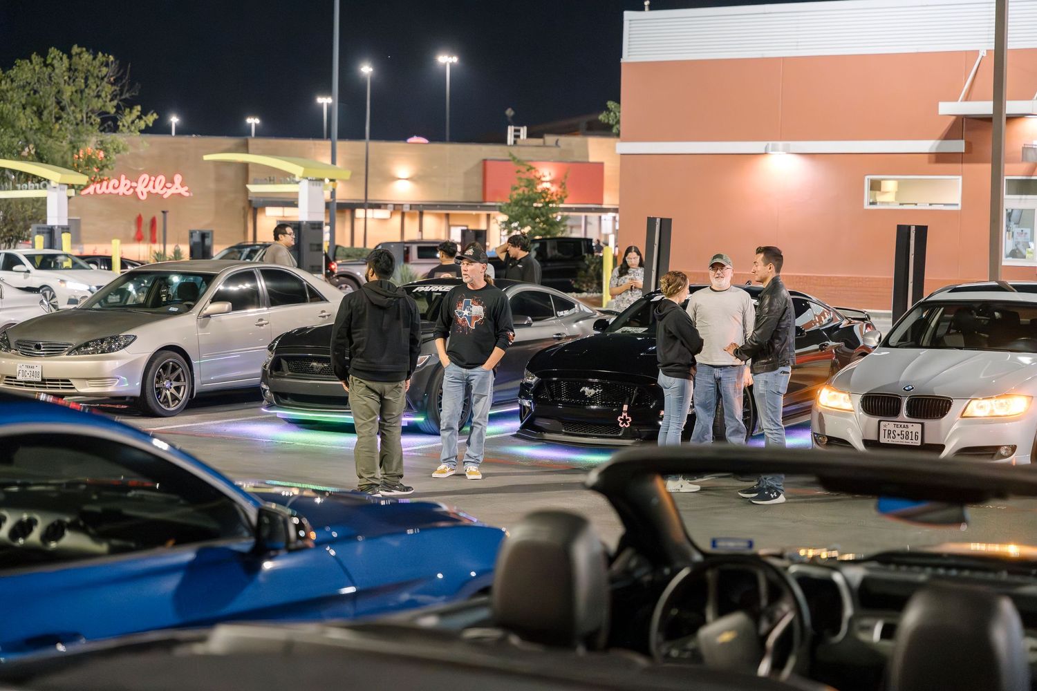 Car enthusiasts gathered around vehicles at a nighttime parking lot meet.