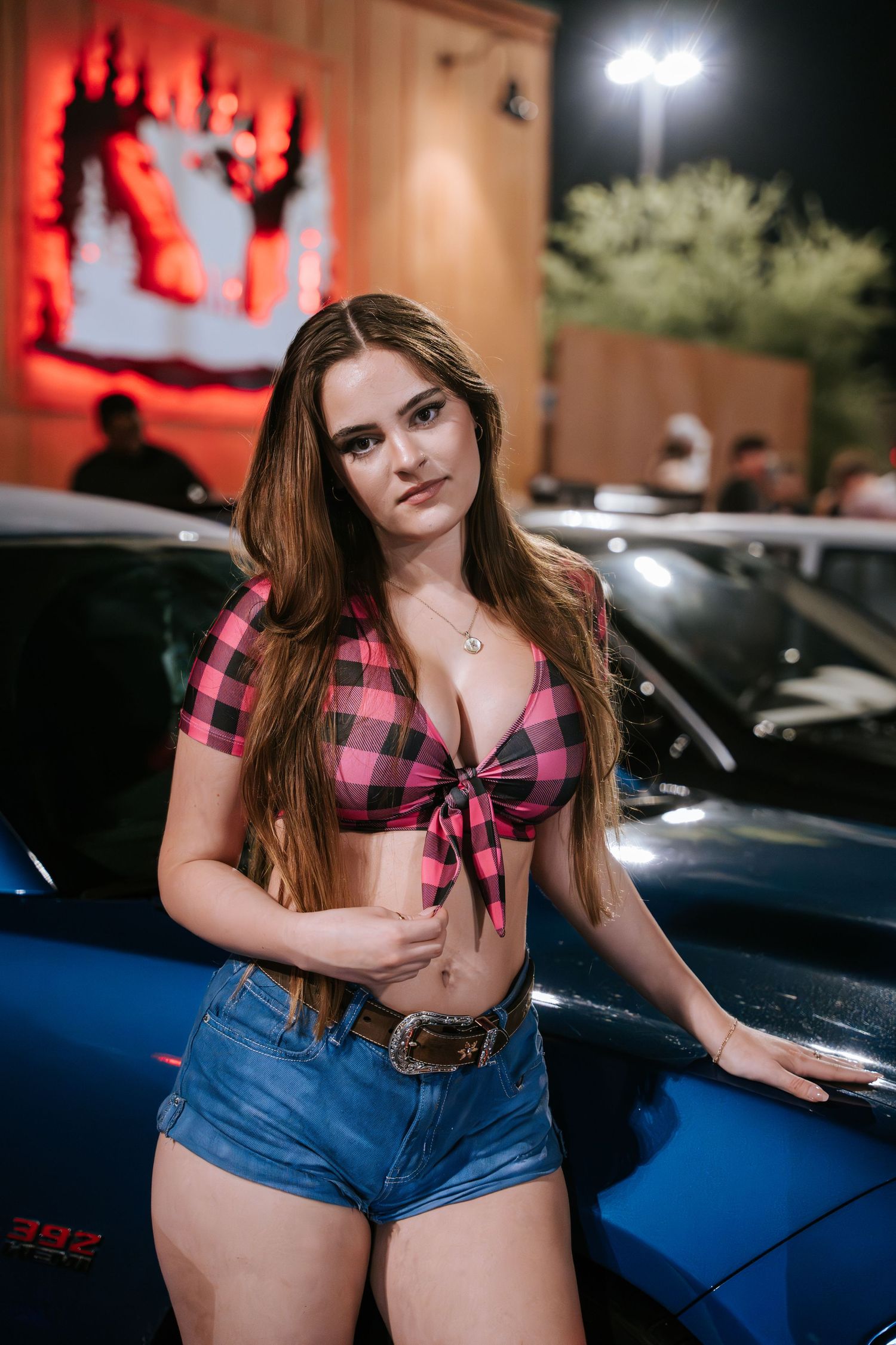 Person in plaid crop top and denim shorts poses next to a blue sports car at night.