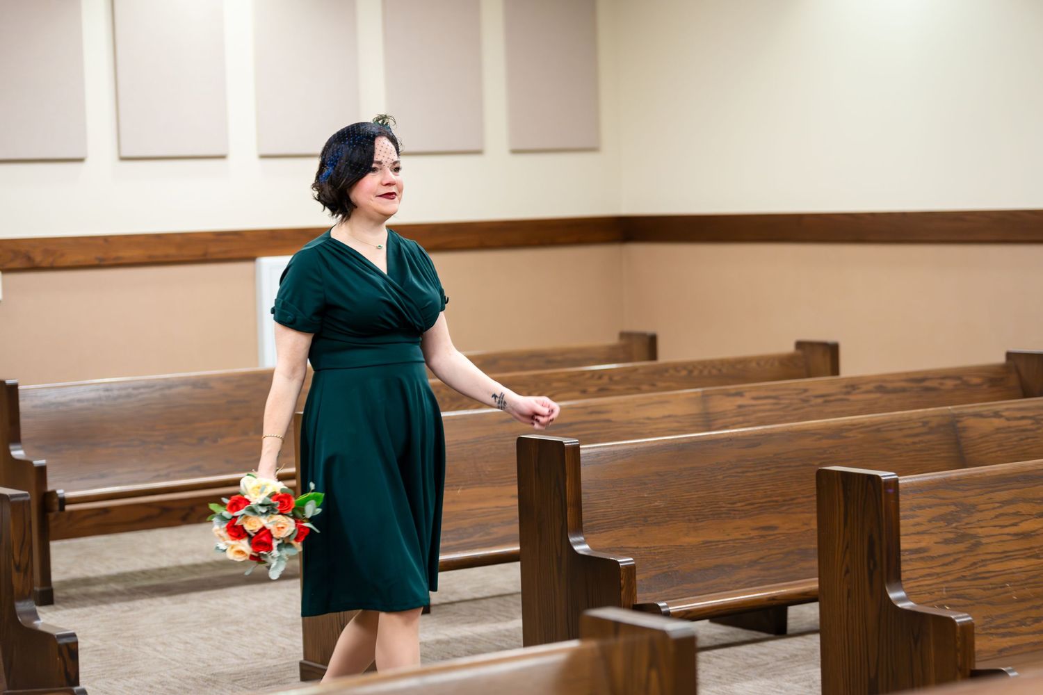 A woman in an emerald green dress stands thoughtfully in a church pew hall holding a bouquet of flowers.