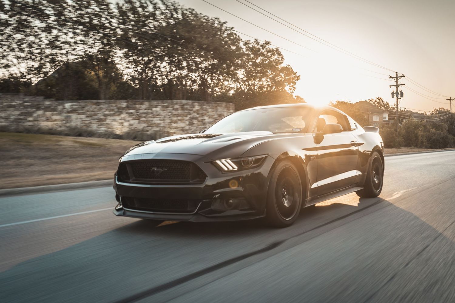 Black Ford Mustang drives on a road with sun flare in the background.