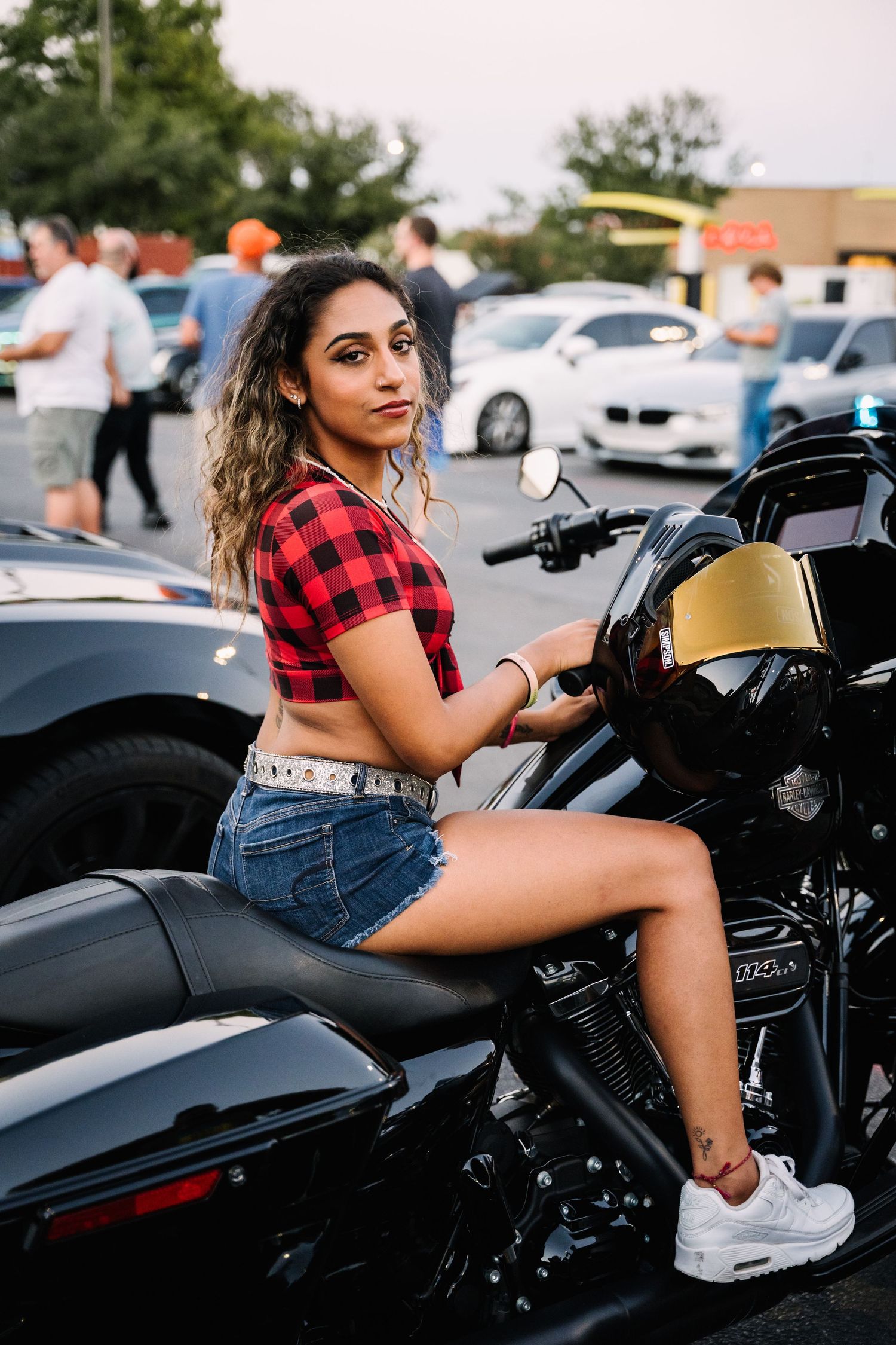 Model in red plaid crop top and denim shorts poses on black motorcycle in parking lot surrounded by cars.