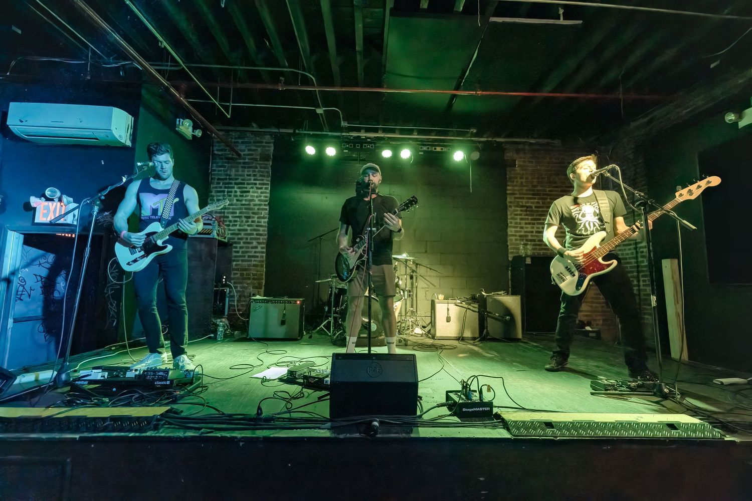 Four-piece band performing on stage with green lighting and exposed brick wall background.