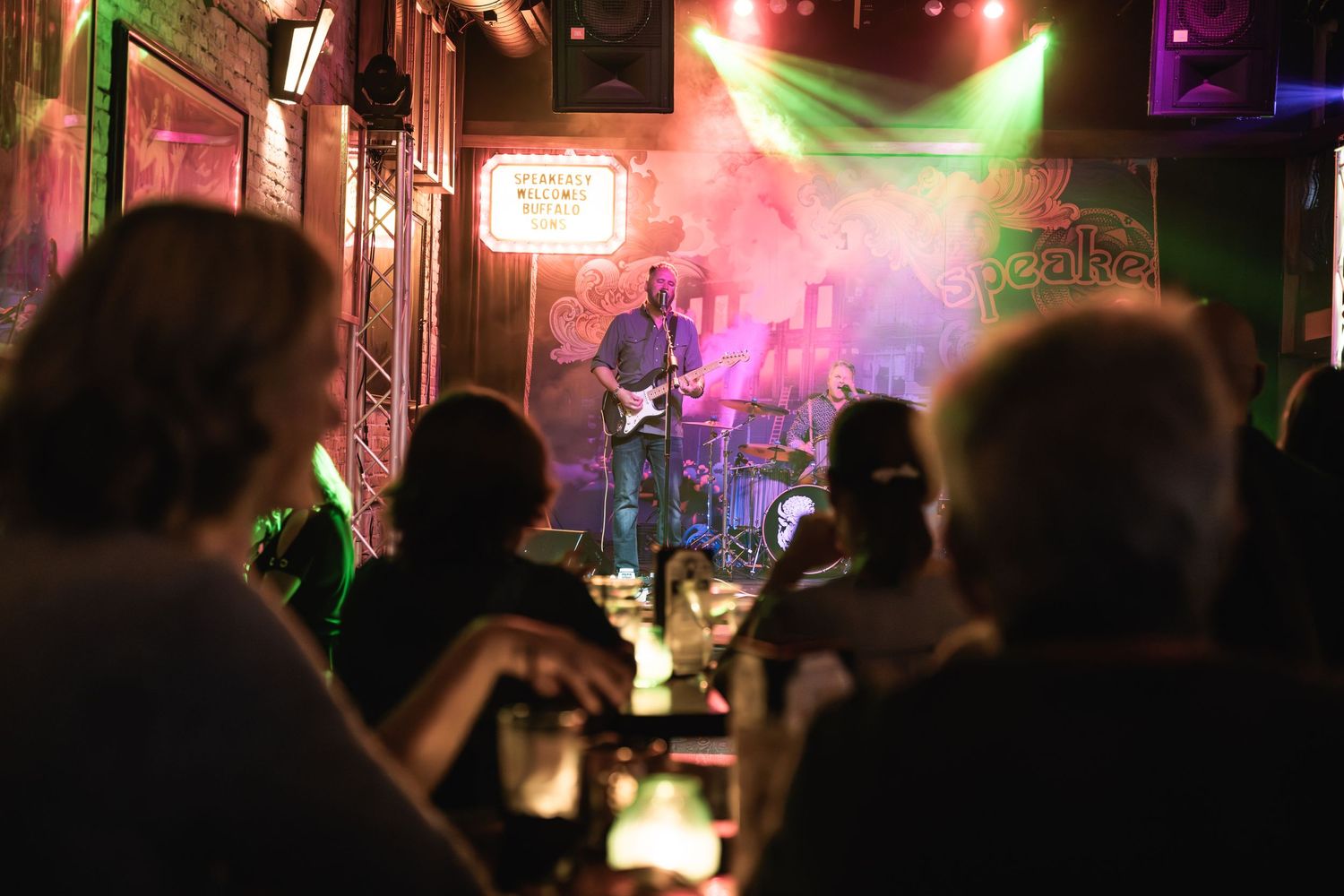 Crowd watches band perform through atmospheric smoke and colored stage lighting.