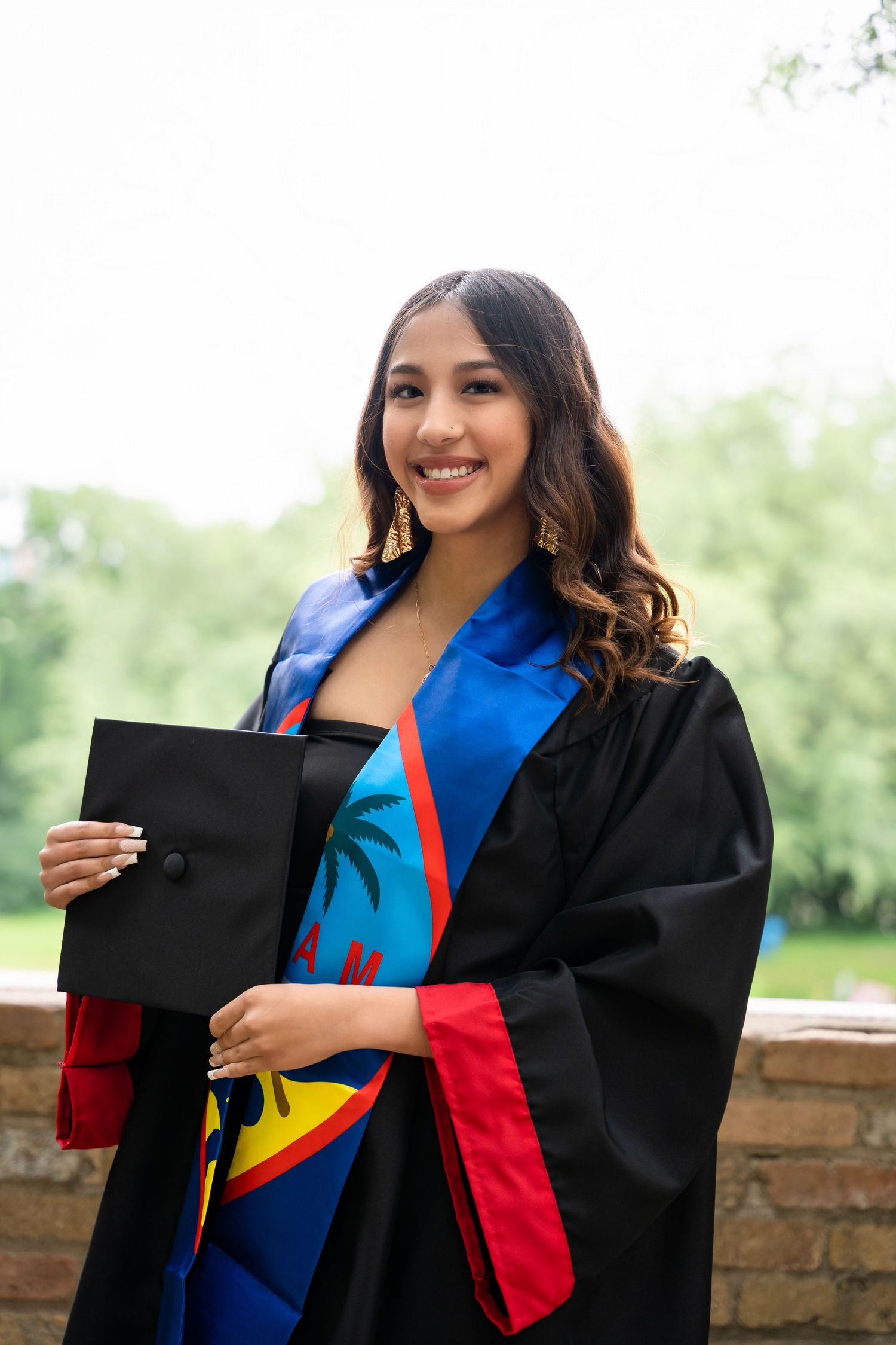 Graduate in ceremonial cap and gown with colorful stole celebrating academic achievement outdoors.