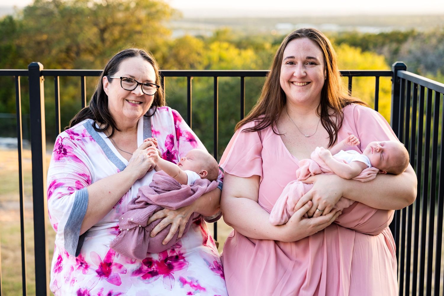 Two women holding newborn babies on outdoor deck during golden hour.