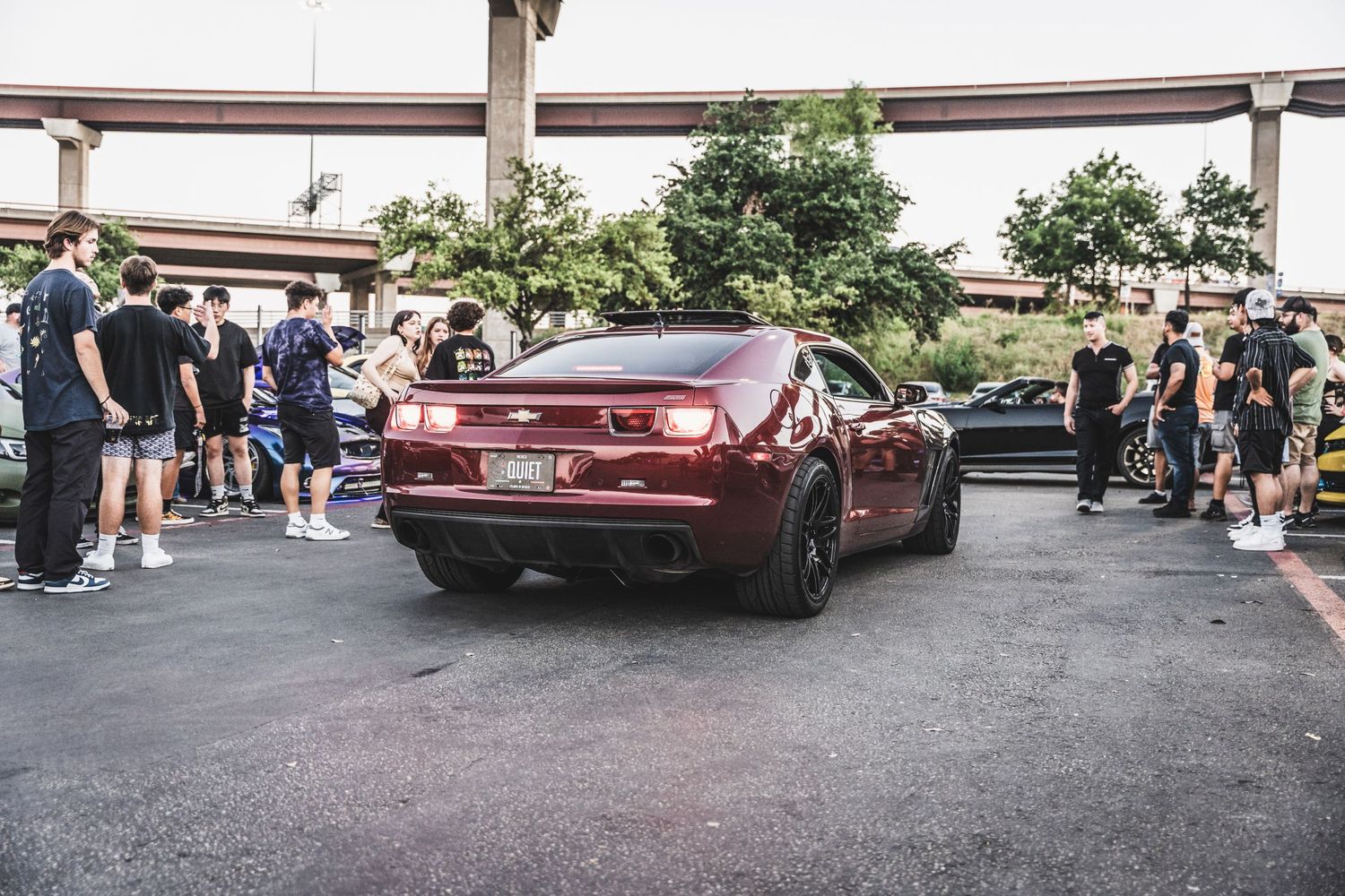 Maroon sports car surrounded by spectators at a car meet under an overpass.