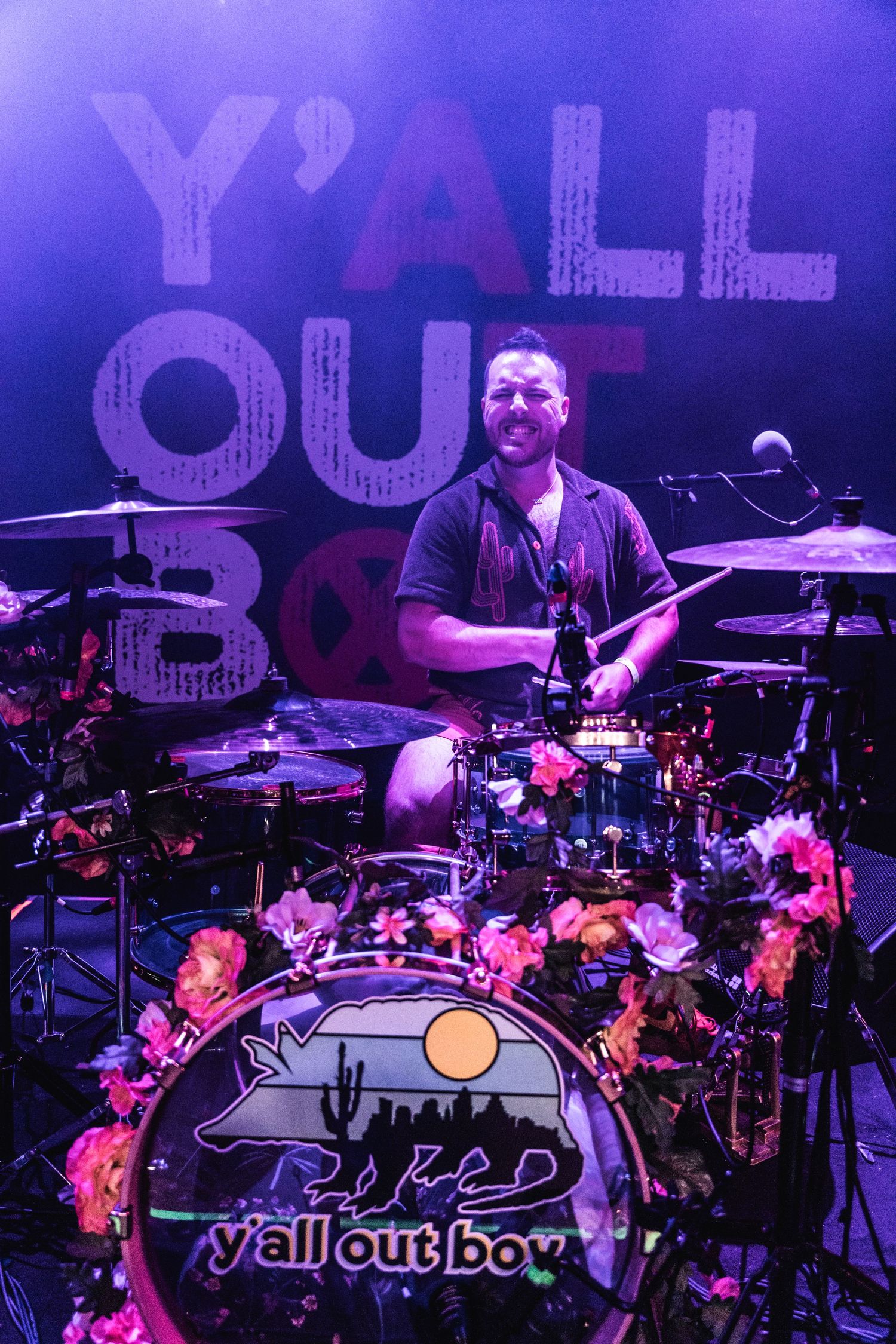 Drummer performs behind a decorated drum kit with Y'all Out Boy text display in background.