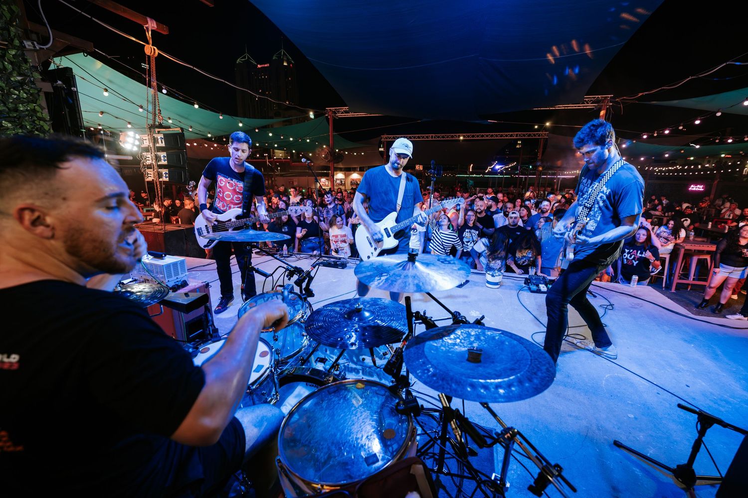 Band performing on stage with drummer and guitarists in blue lighting at live music venue.
