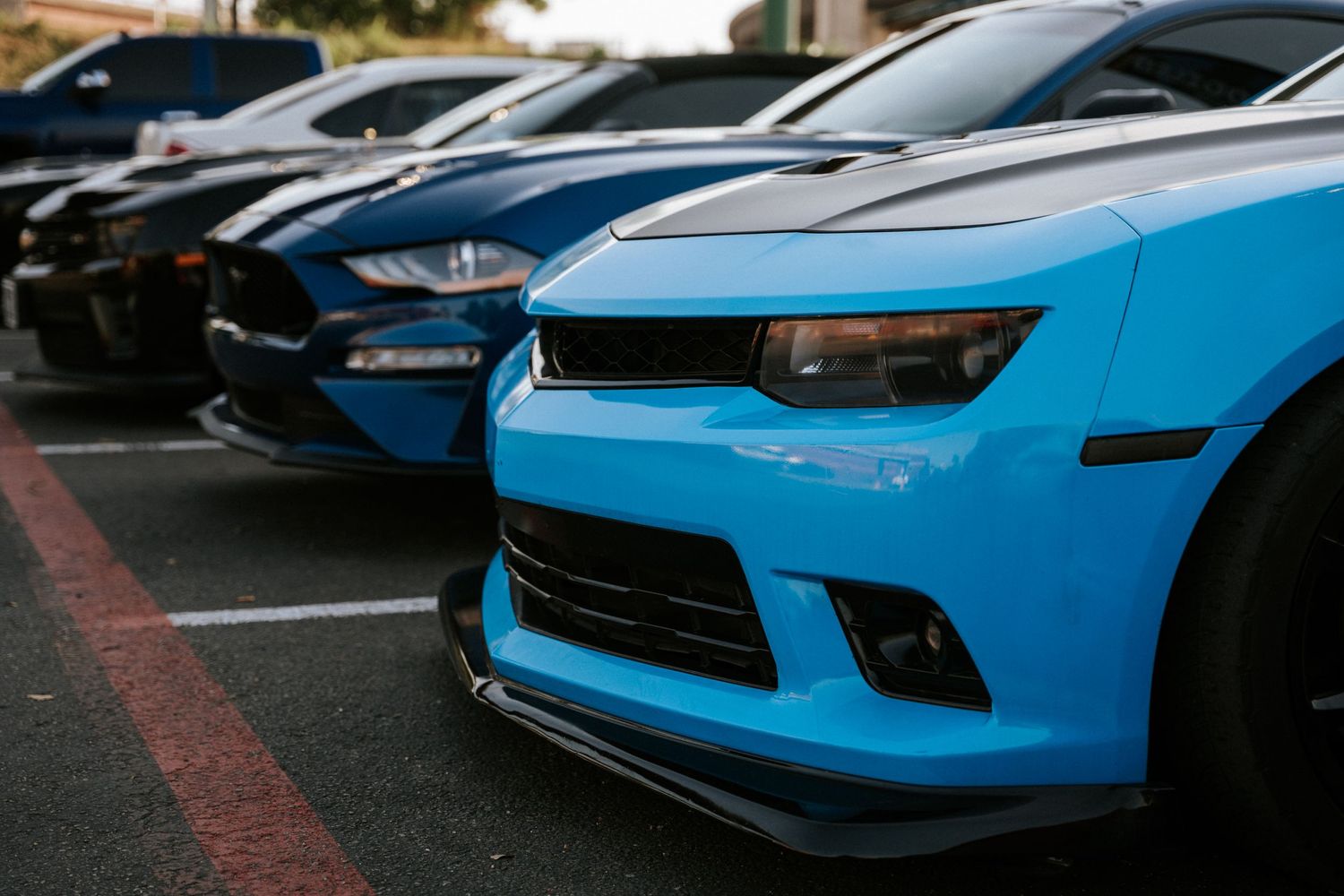 Row of modified sports cars including a bright blue Camaro and dark colored muscle cars in a parking lot.
