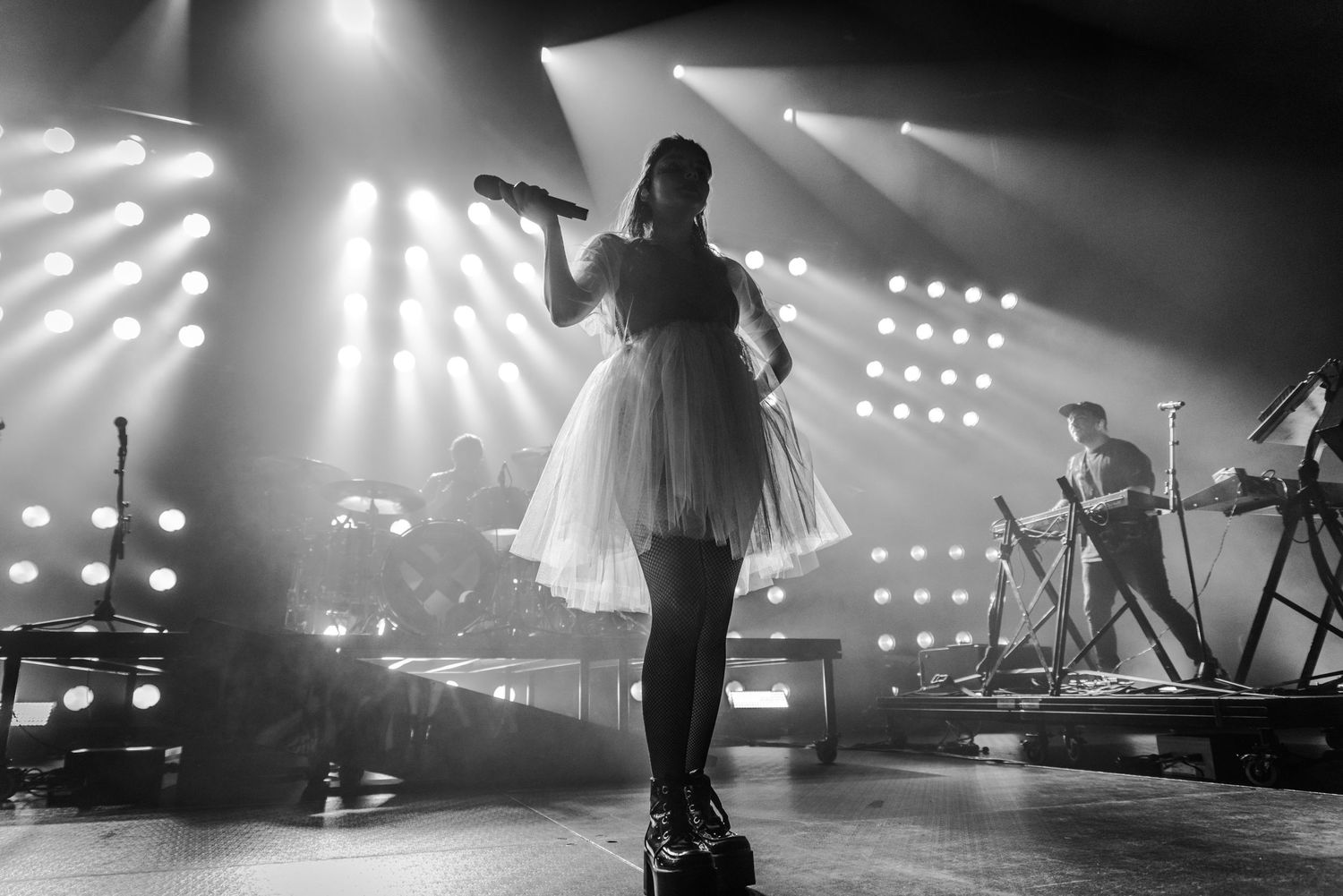 Silhouetted concert performer in dress stands elevated on stage with dramatic backlighting.