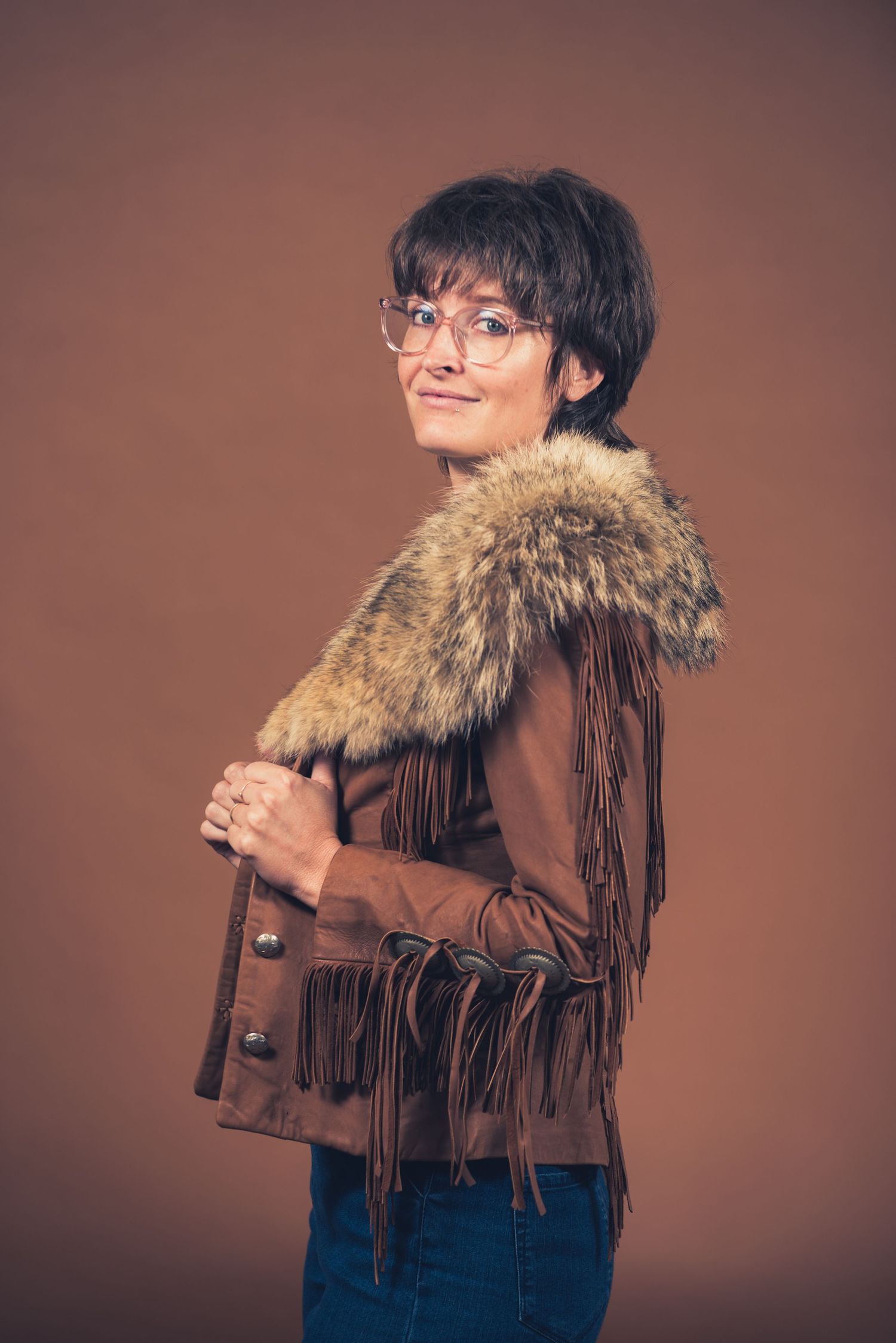Person wearing vintage-style fringed leather jacket with fur collar and glasses in studio setting.