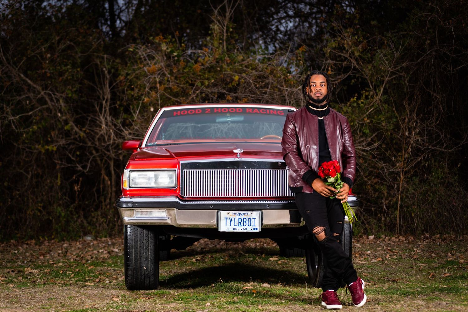 Person in casual wear poses with a vintage red car against a wooded backdrop.