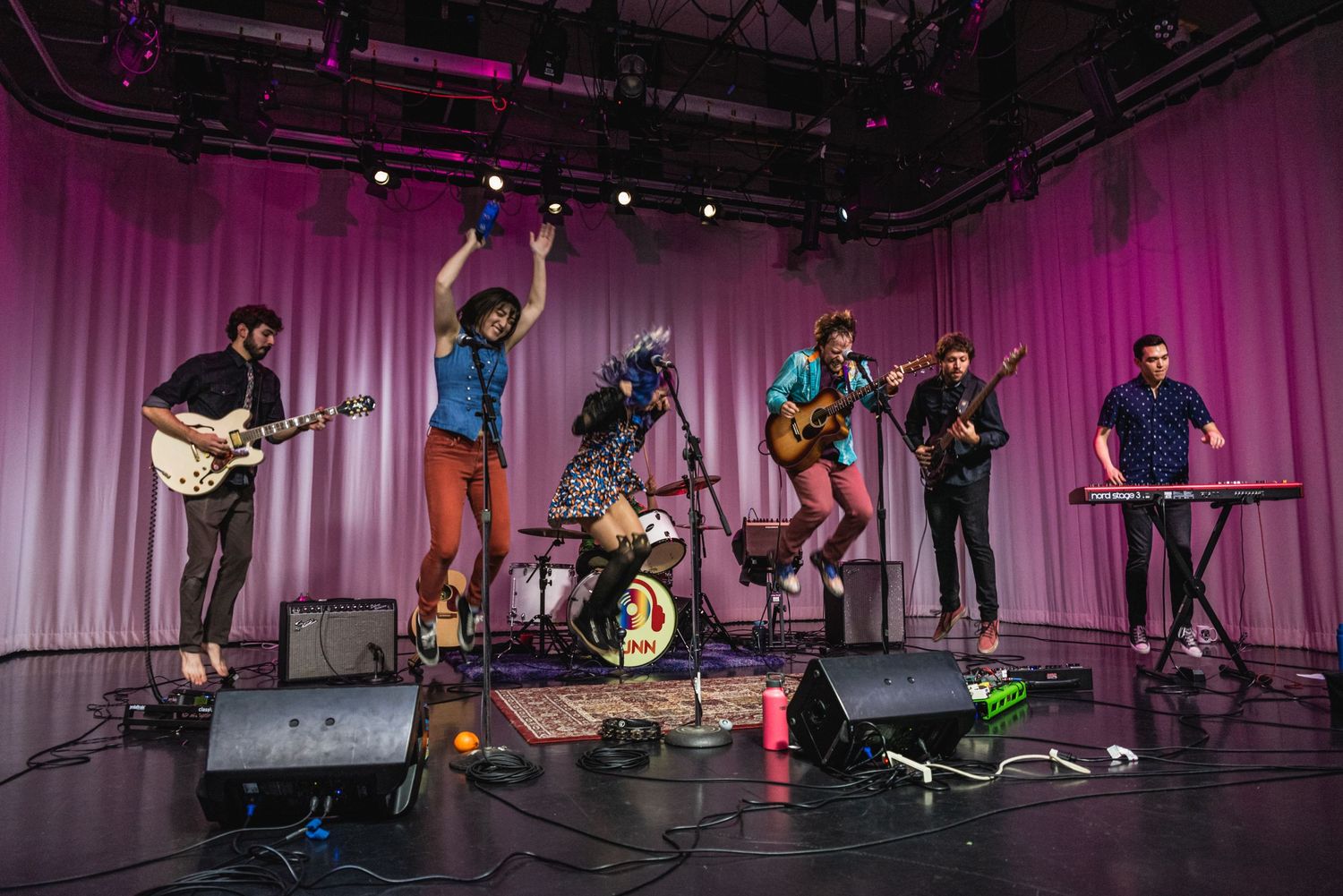 Musicians perform energetically on stage with instruments against pink curtain backdrop during live show.