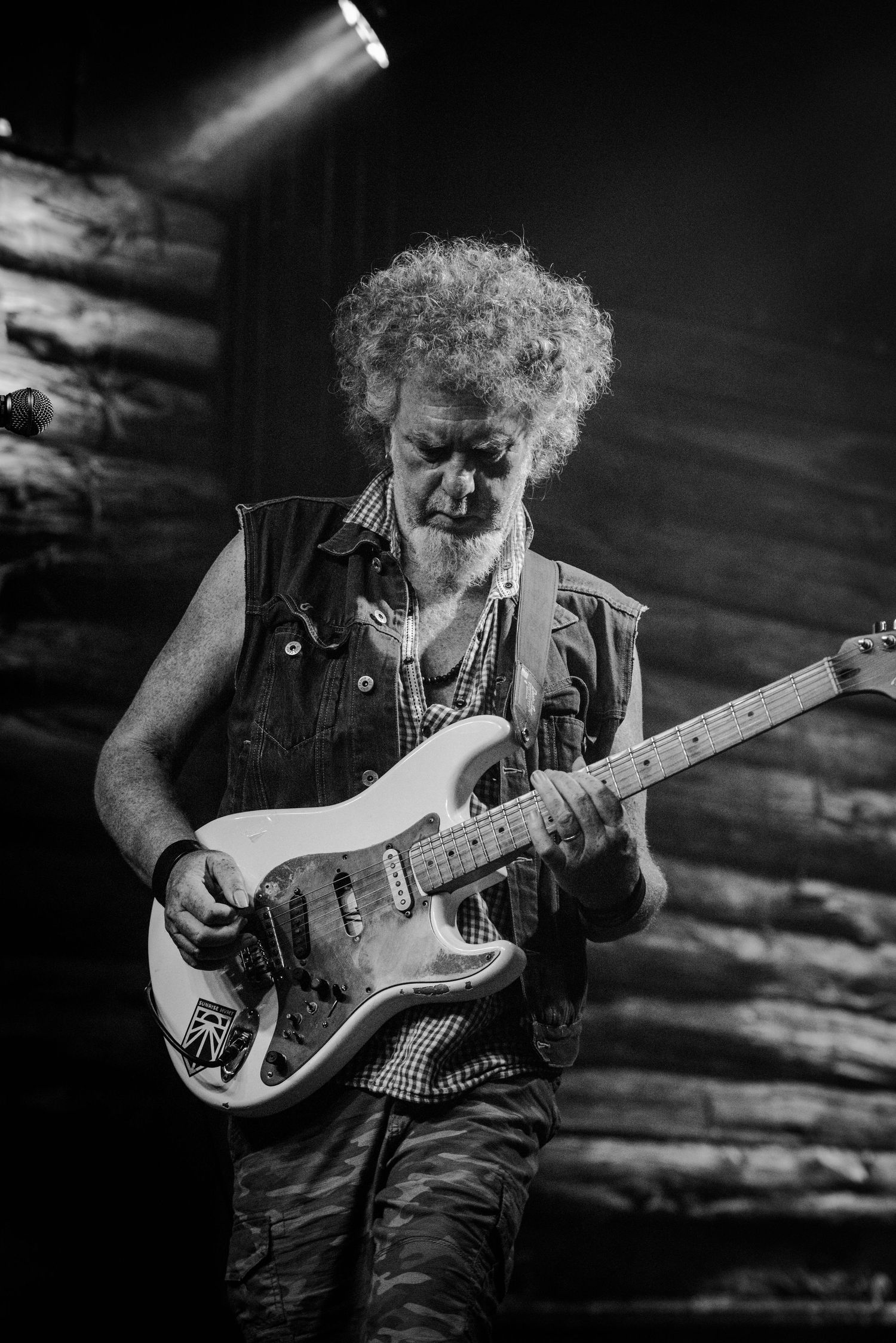 Guitarist with curly hair plays electric guitar while wearing a denim vest during dramatic stage performance.