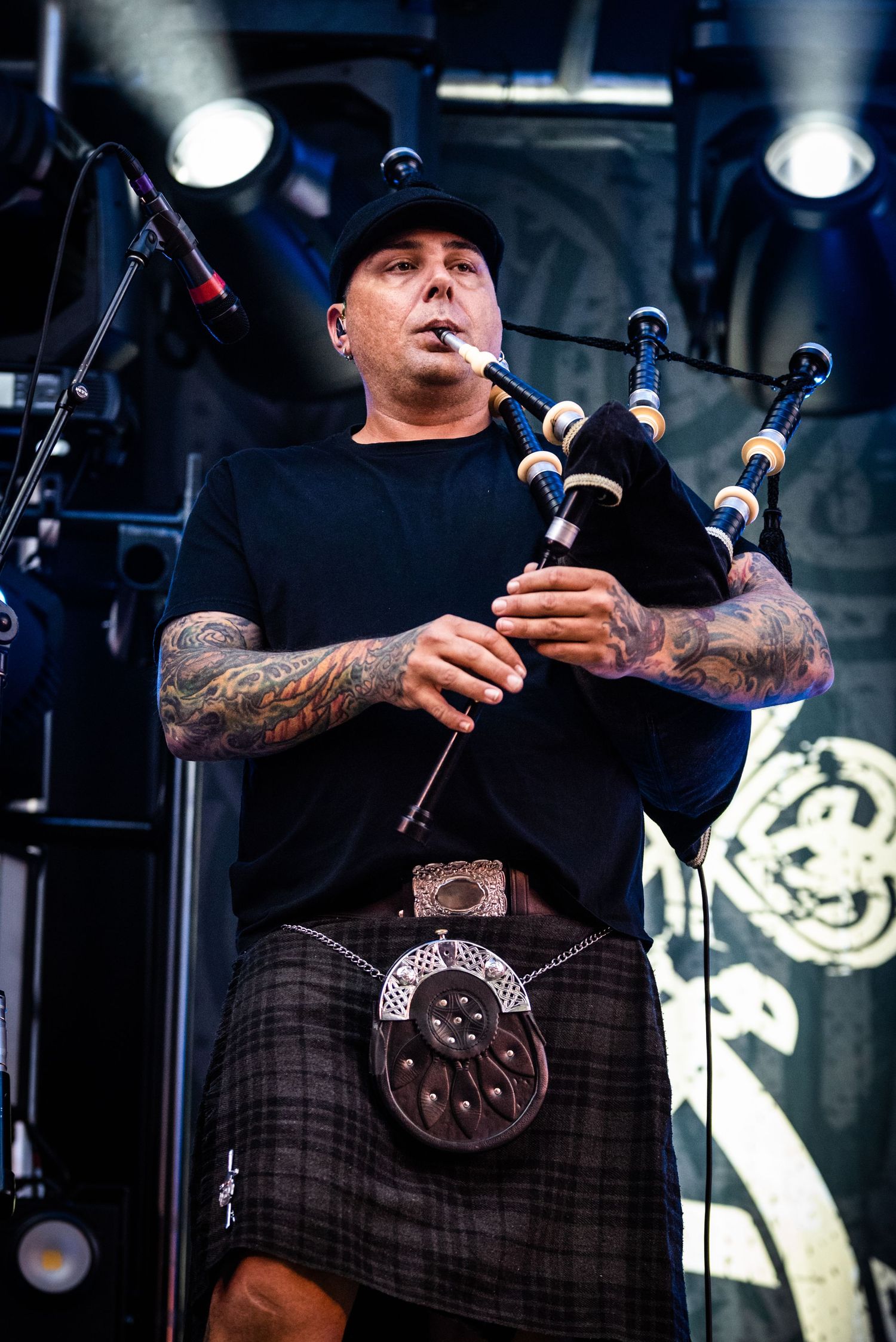 Musician in black shirt and kilt plays bagpipes on stage with tattooed arms.