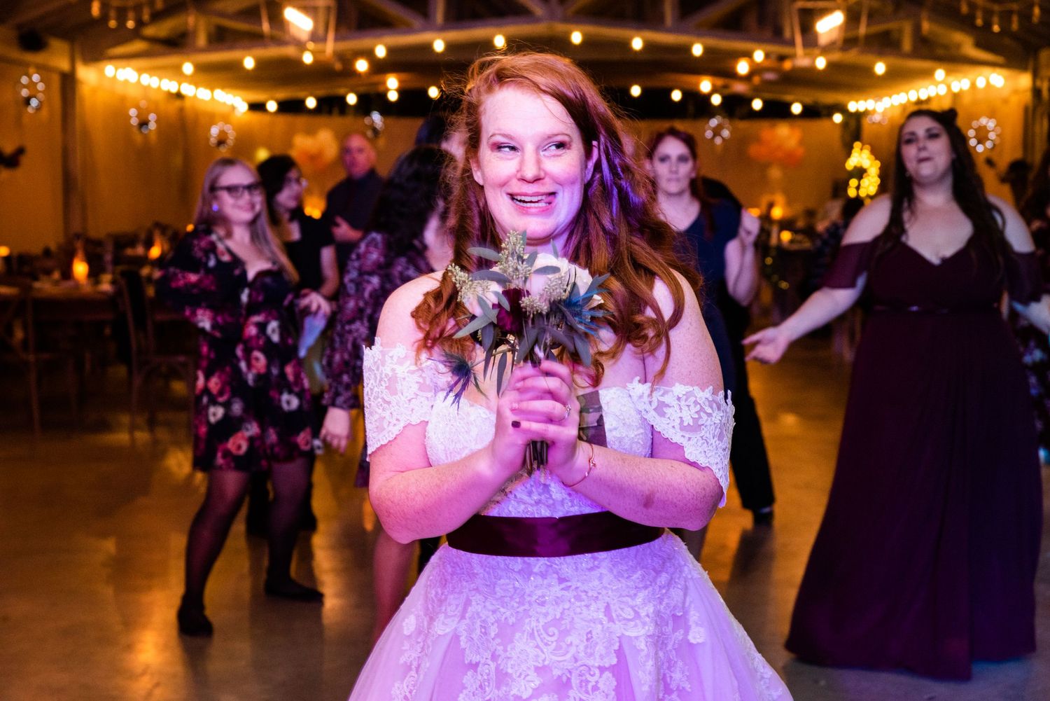 Bride in a lace wedding dress holds bouquet while dancing under string lights.
