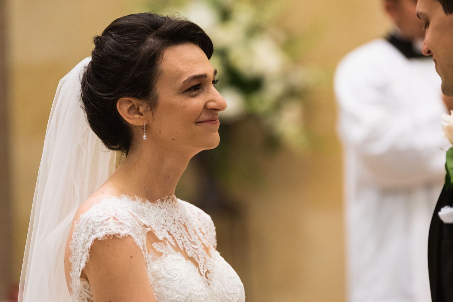 Bride in lace wedding gown smiling during ceremony with veil and updo hairstyle.