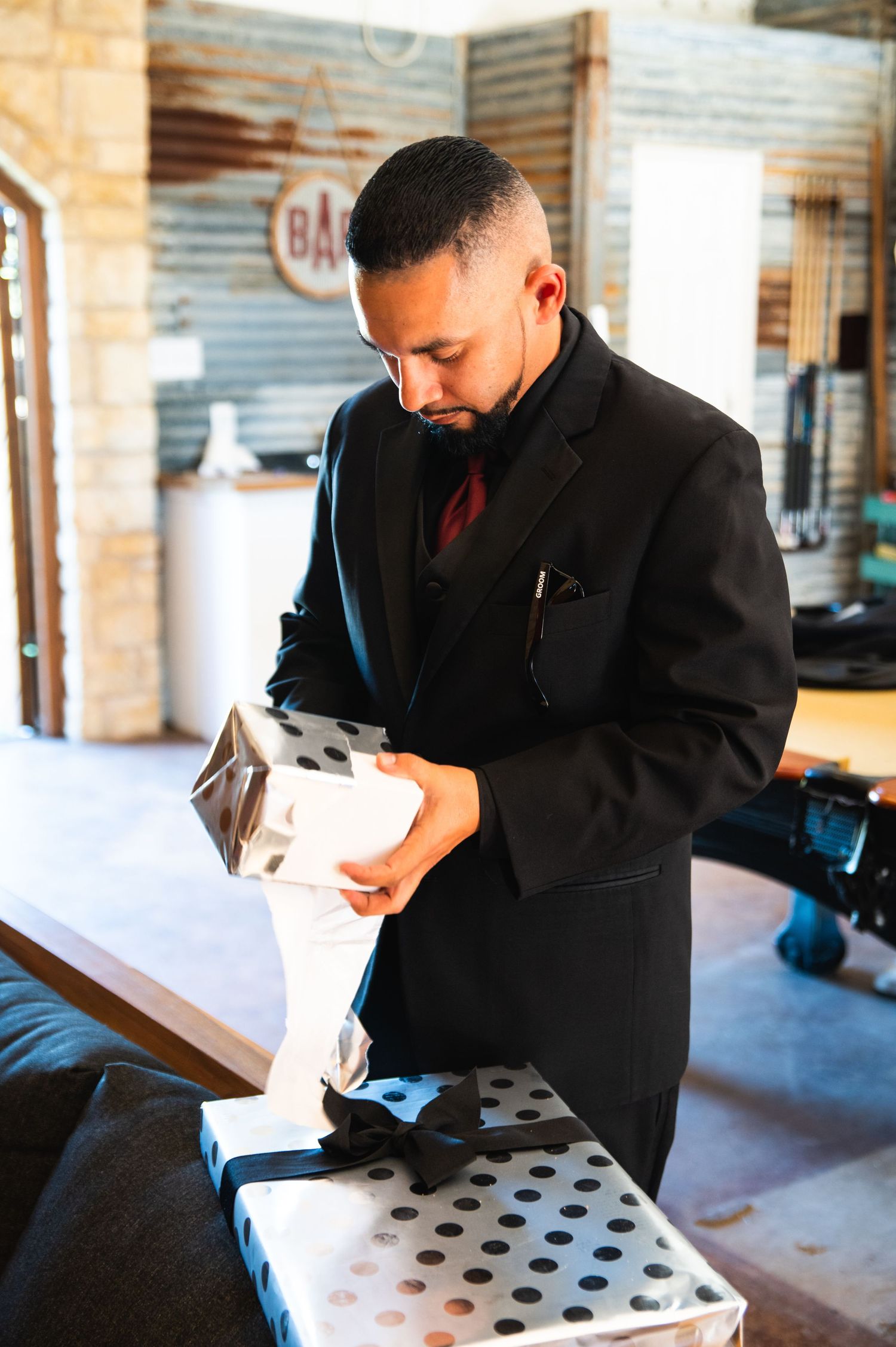 A person in a black suit examines an unwrapped gift in a rustic room.