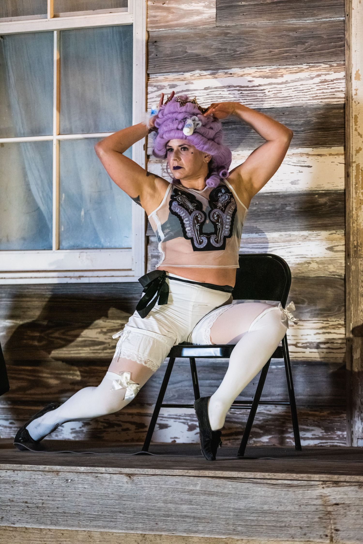 Performer in metallic crop top and white pants poses dramatically on wooden porch.