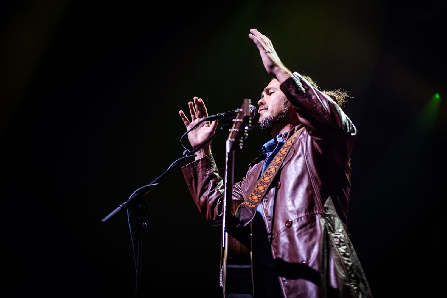 Performer in brown leather jacket sings emotionally with raised hand during concert performance.