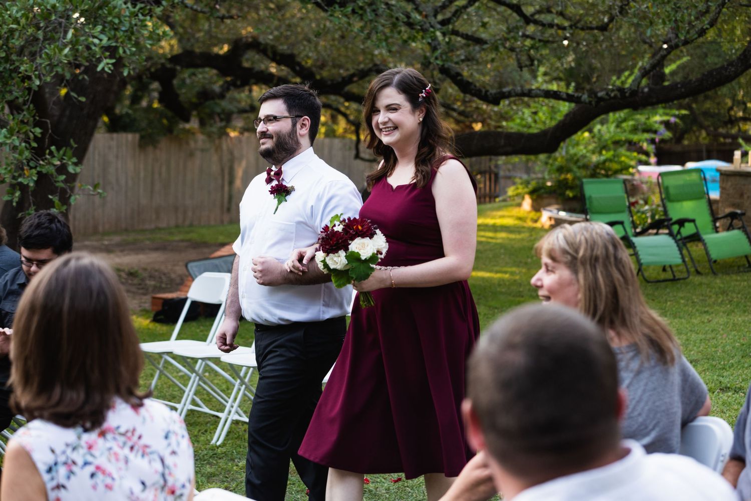 Wedding attendees in formal attire gather at an outdoor reception with white chairs.