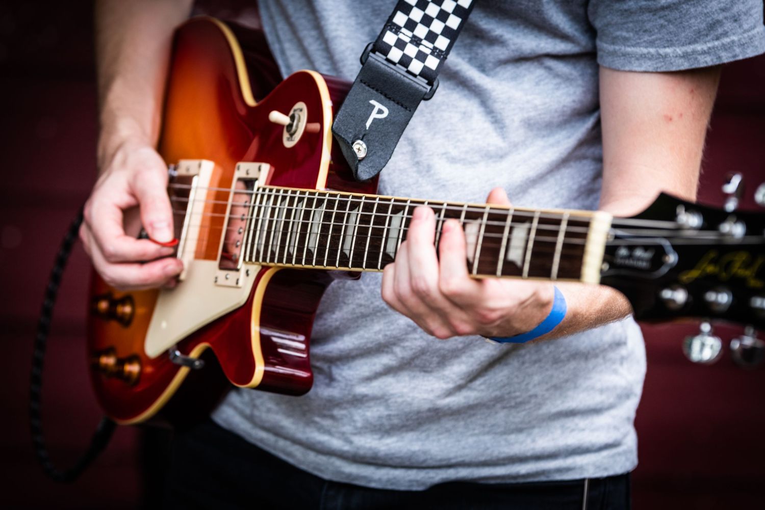 Close-up shot of hands playing red electric guitar with checkerboard guitar strap.