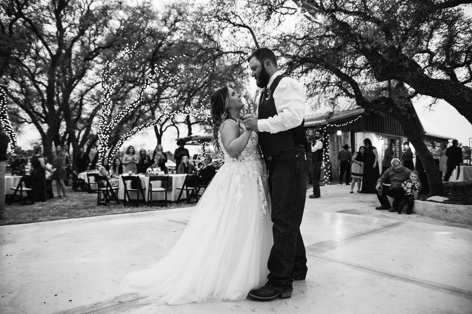 Black and white photo of newlyweds dancing on an outdoor patio with guests seated at tables nearby.