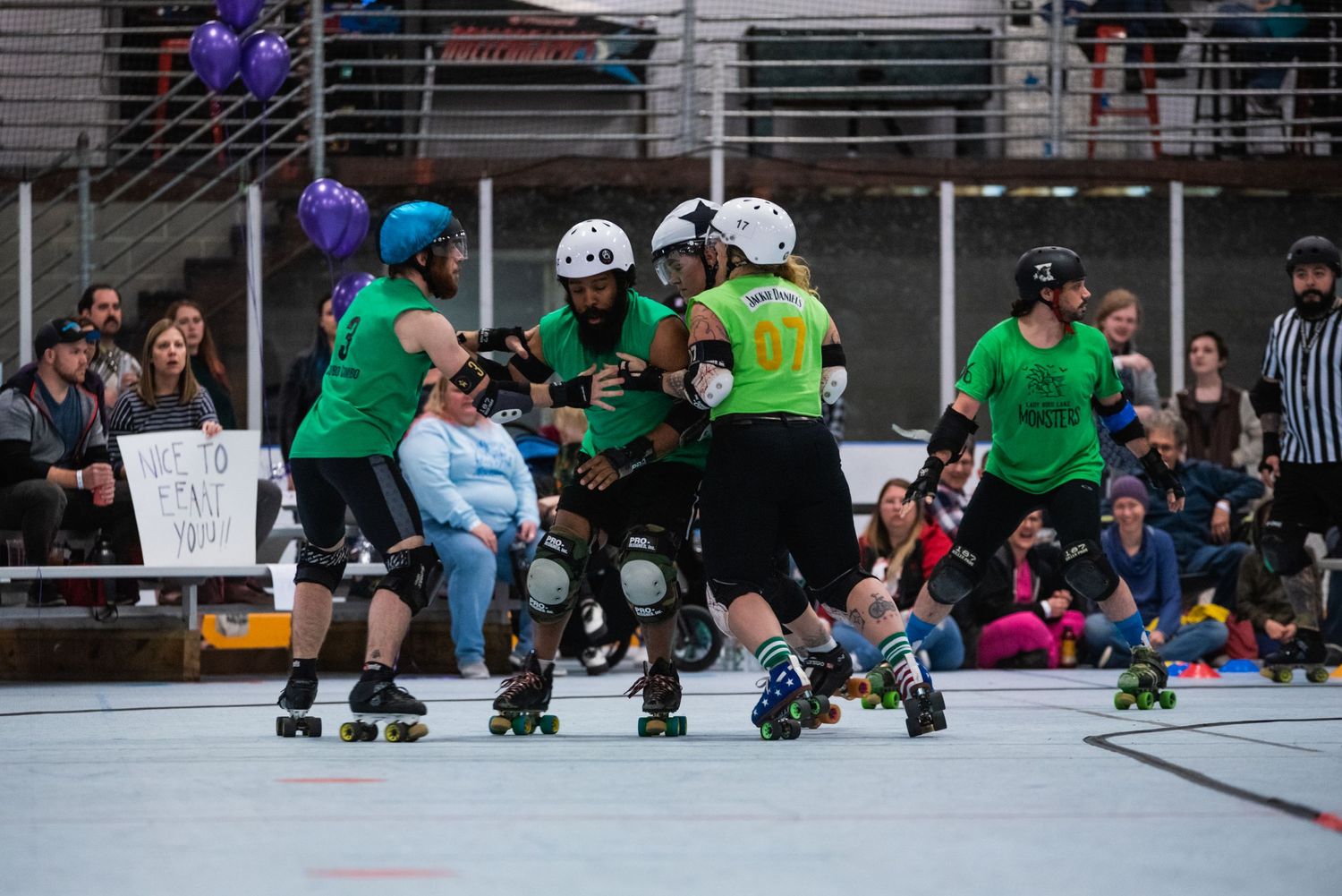 Roller derby team in green uniforms competing on indoor track with spectators watching.