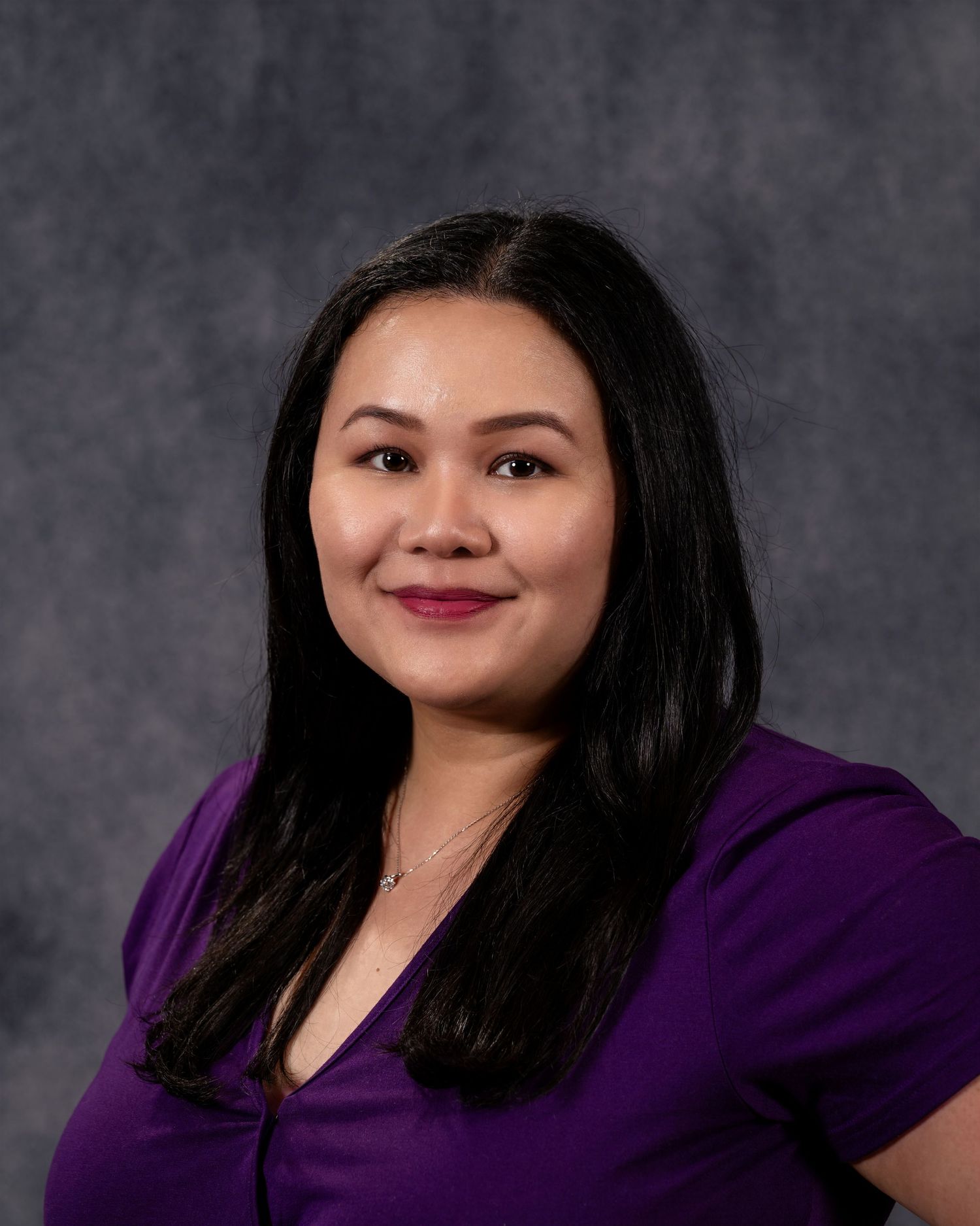 Professional headshot of a smiling woman with long black hair wearing a purple top against a gray background.