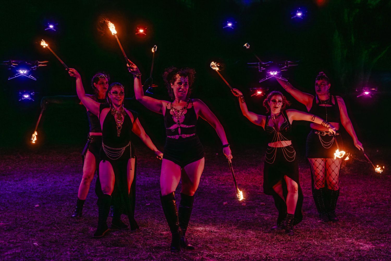 Five fire dancers perform at night, wielding flaming torches under purple and blue lights in dramatic costumes.