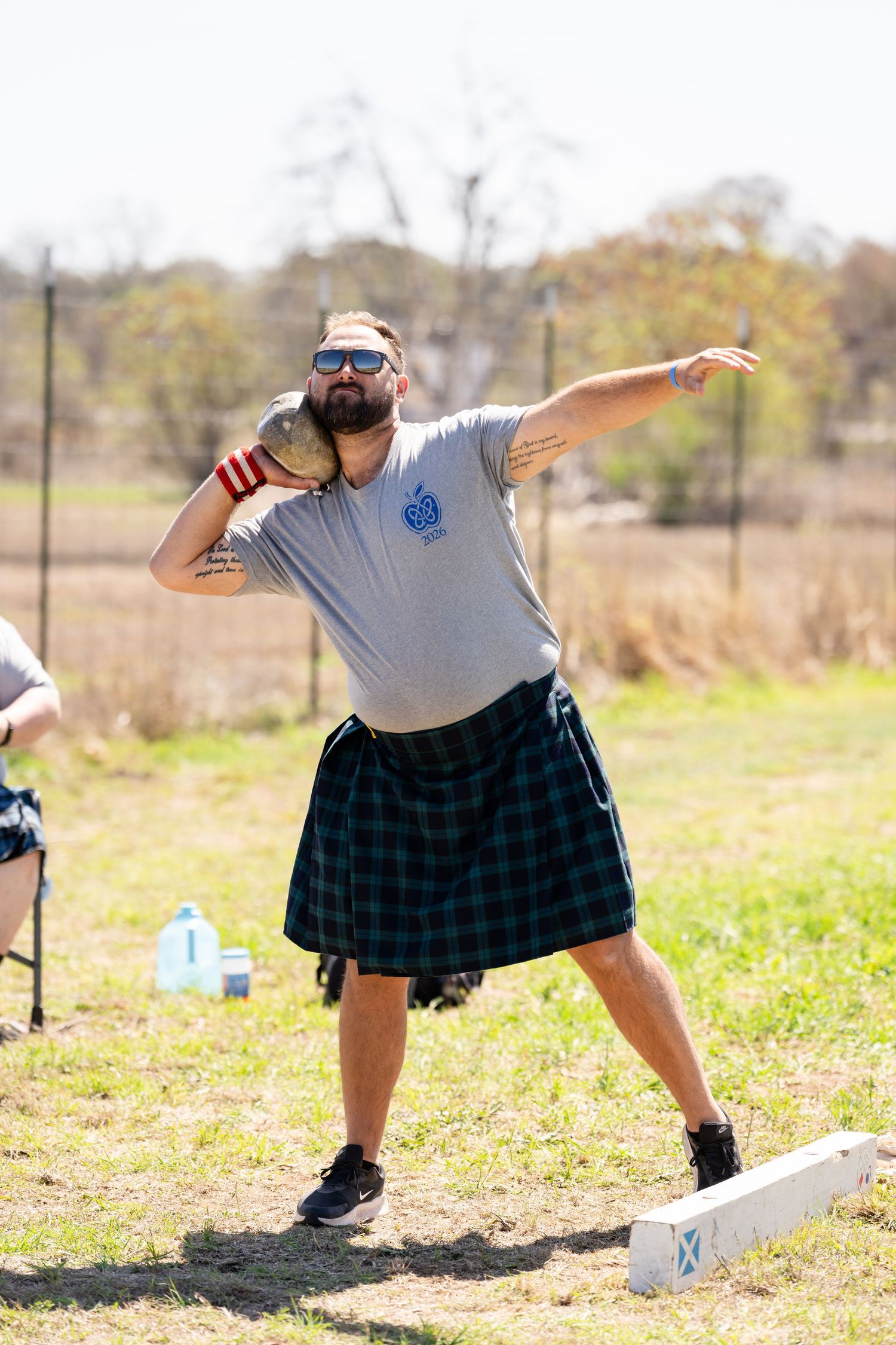 Man in kilt throwing shot put at Highland Games competition outdoors.