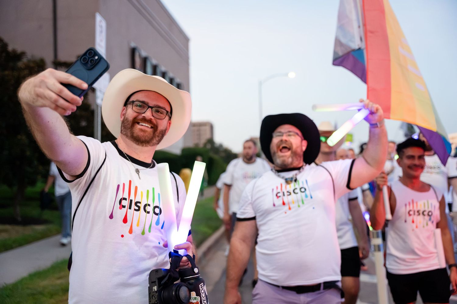 Two men in Cisco t-shirts and cowboy hats take a selfie at a Pride parade, holding rainbow flags and glowing sticks.