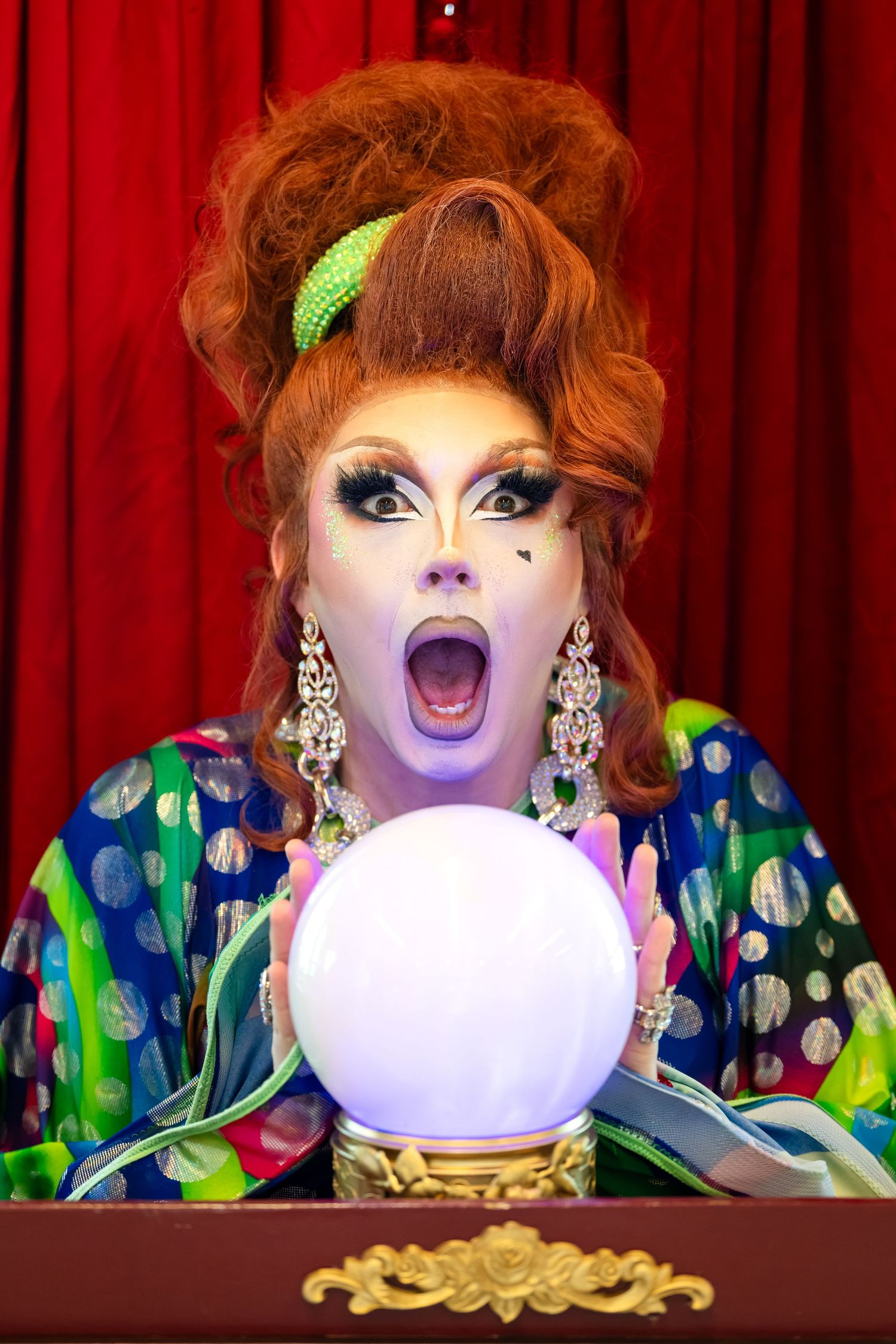 Drag queen in colorful polka dot costume gazes into glowing crystal ball with dramatic expression on stage.