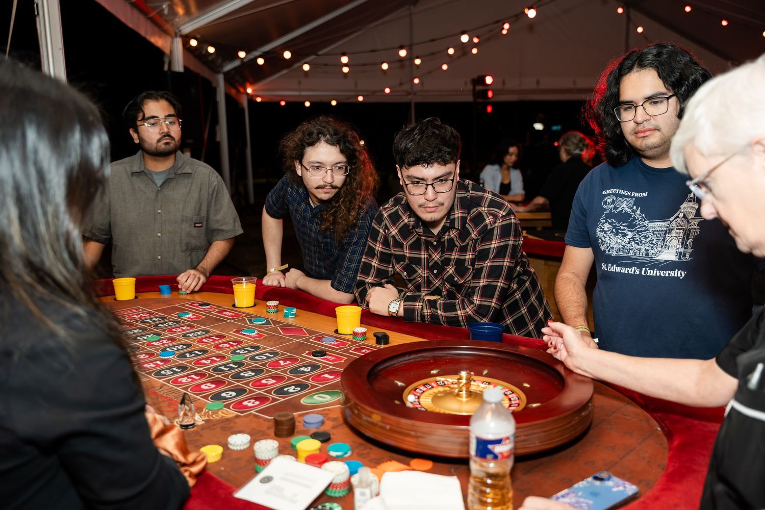 A group of people playing roulette and gambling at a casino table with red felt and colorful betting chips.