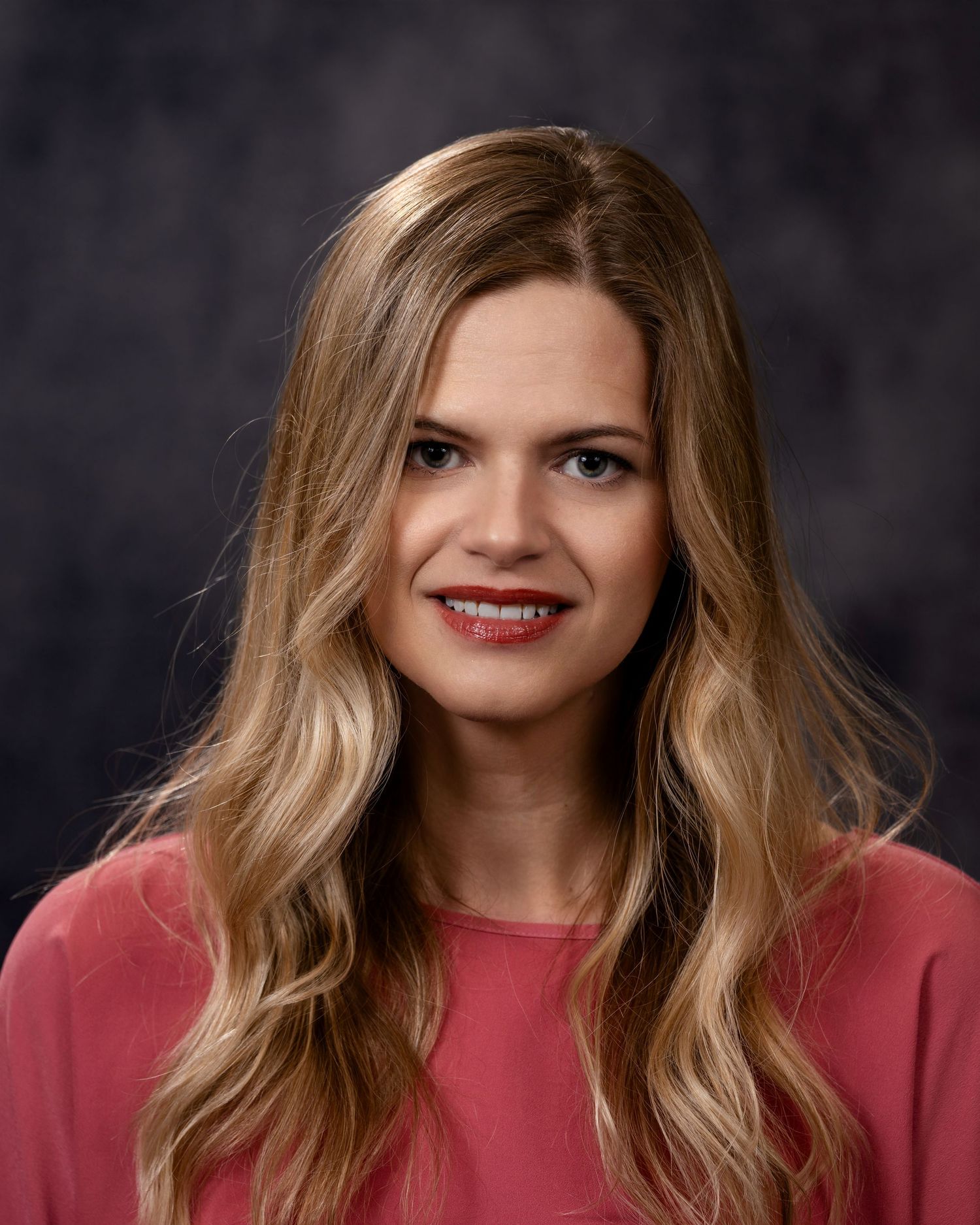 A person wearing a coral-colored sweater smiles warmly against a dark studio background.