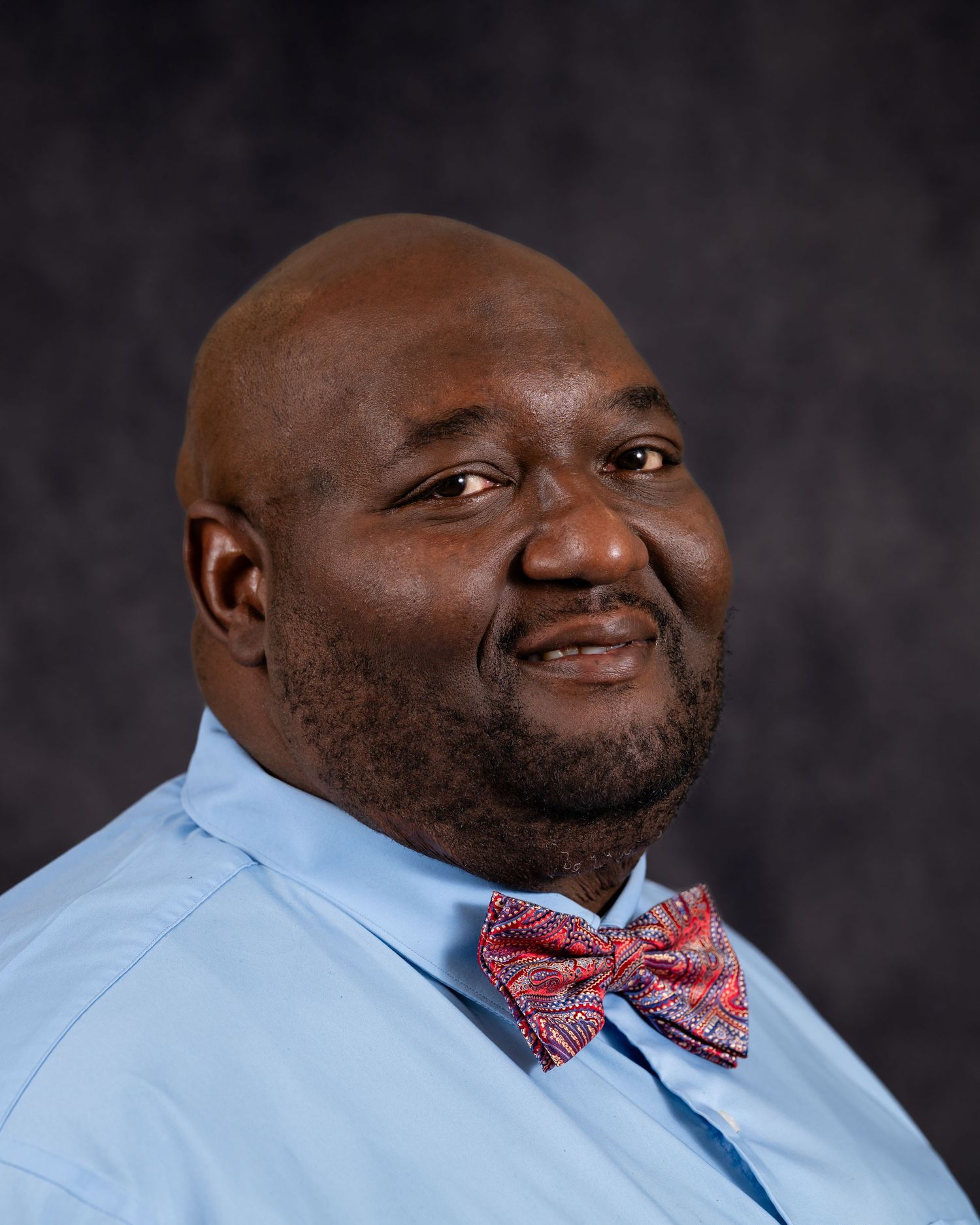 Professional headshot portrait of a person wearing a light blue dress shirt and red patterned bow tie against a dark background.