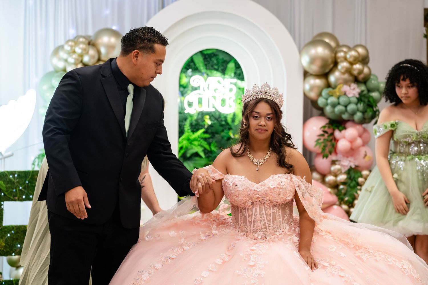 A young woman in a pink quinceañera gown wearing a tiara dances with a man in a black suit at her celebration.